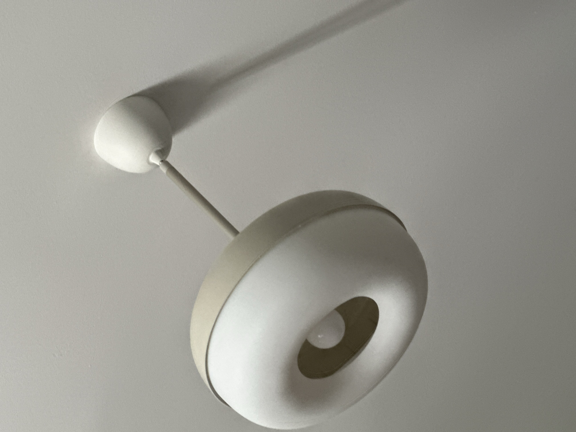 A ceiling light fixture is shown, suspended from a plain white ceiling. The fixture has a beige outer ring and a white, toroidal-shaped diffuser. A lightbulb is visible within the central opening of the diffuser. A shadow from the fixture is cast onto the ceiling to the right. The image is taken from a low angle, looking upwards. The environment appears to be indoors, likely a residential or commercial space. The specific location in Fethiye, Turkey, cannot be determined from the visual information provided.