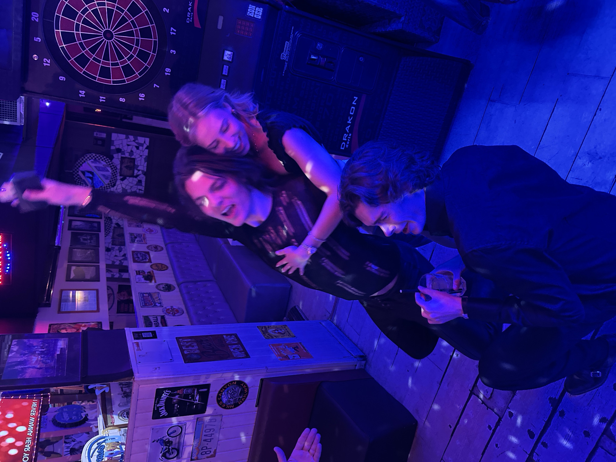 A horizontal eye-level shot shows three individuals, two women and a man, in a bar or club, illuminated by blue lighting. In the background is a dartboard and electronic dart machine. The younger woman is hugging the middle-aged woman from behind. The man is crouching on the right with a glass. There are various signs and posters on the walls, including ones for Jack Daniel's and Canyon Railway. There is a sign indicating "OPEN ONE." The location is Madrid, Spain, as indicated by the license plate "8P" referencing Madrid.