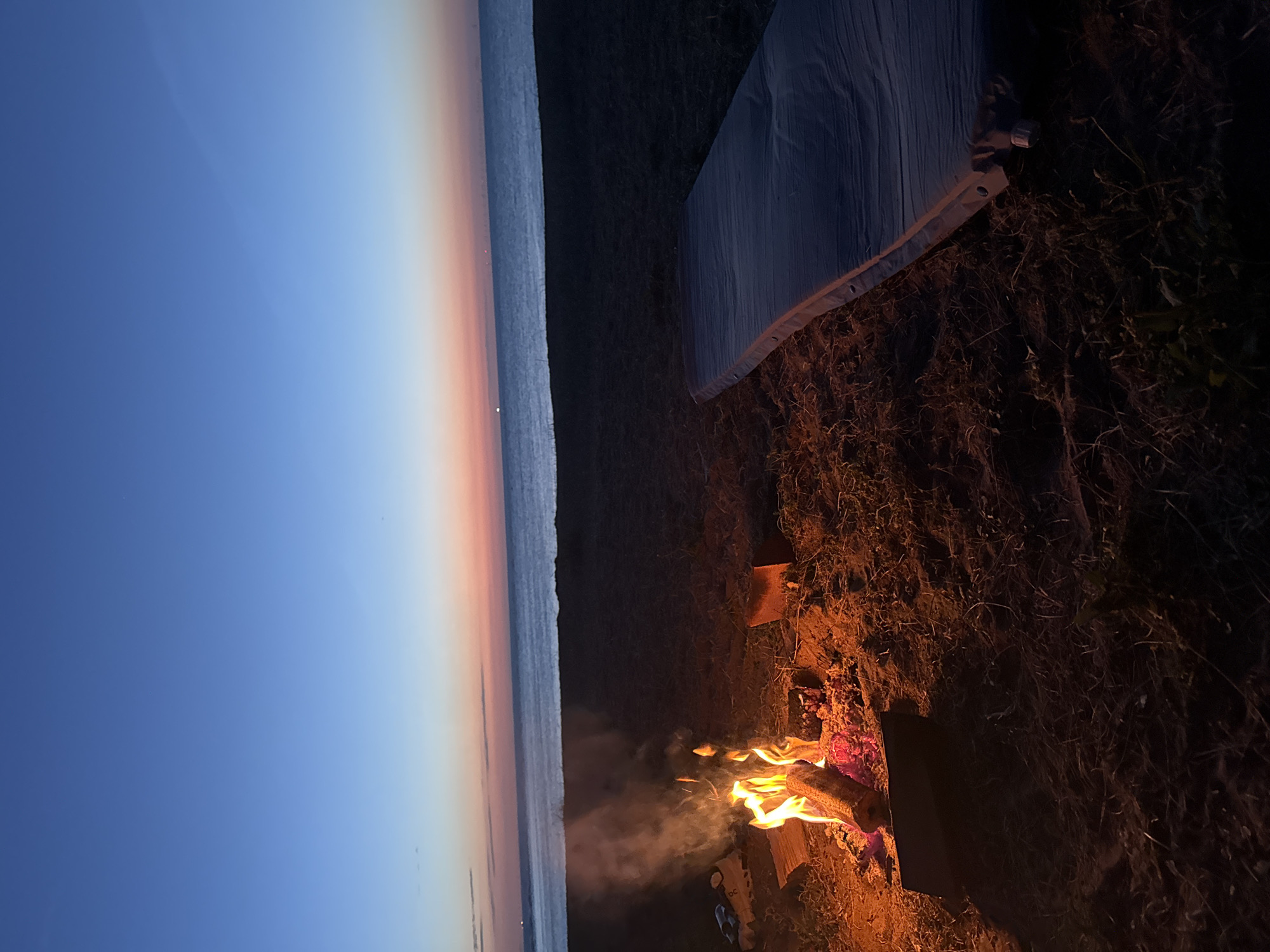 A nighttime scene of a beach campsite on the coast of Thanet, United Kingdom. The sky transitions from a deep blue overhead to hues of orange and pink near the horizon, indicating twilight or dawn. A small bonfire burns brightly in the foreground, with logs smoldering and flames reaching upwards, emitting a visible trail of smoke. To the right of the fire, a deflated sleeping mat lies on the sandy ground, partially covered with dried grass and twigs. The sea is visible in the background, appearing calm and reflecting the faint light of the sky. The overall atmosphere suggests a solitary and peaceful camping experience.
