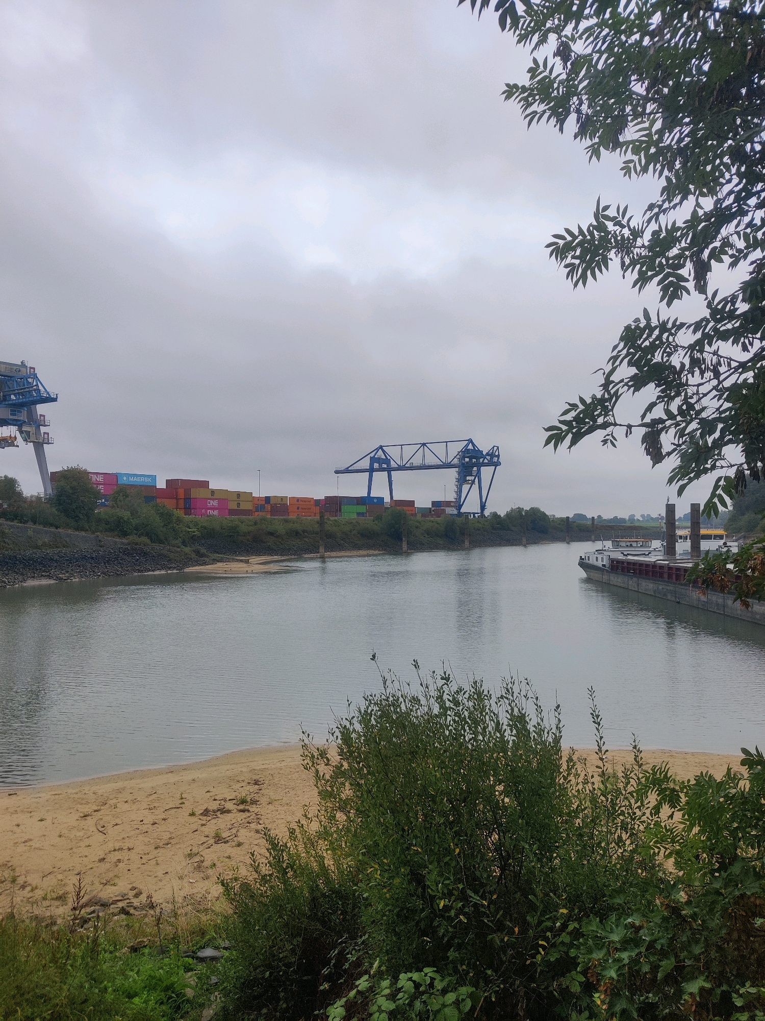 The image shows a wide, calm river flowing through a port area. On the left bank, a large blue gantry crane is visible, along with stacks of colorful shipping containers. Further down the river, another, smaller blue gantry crane stands. The right bank of the river features a barge docked. The sky is overcast and gray. The foreground consists of a sandy bank with some vegetation and trees. The scene is consistent with the port of Emmerich on the Rhine in Germany, a significant inland port. No people are visible in the image.