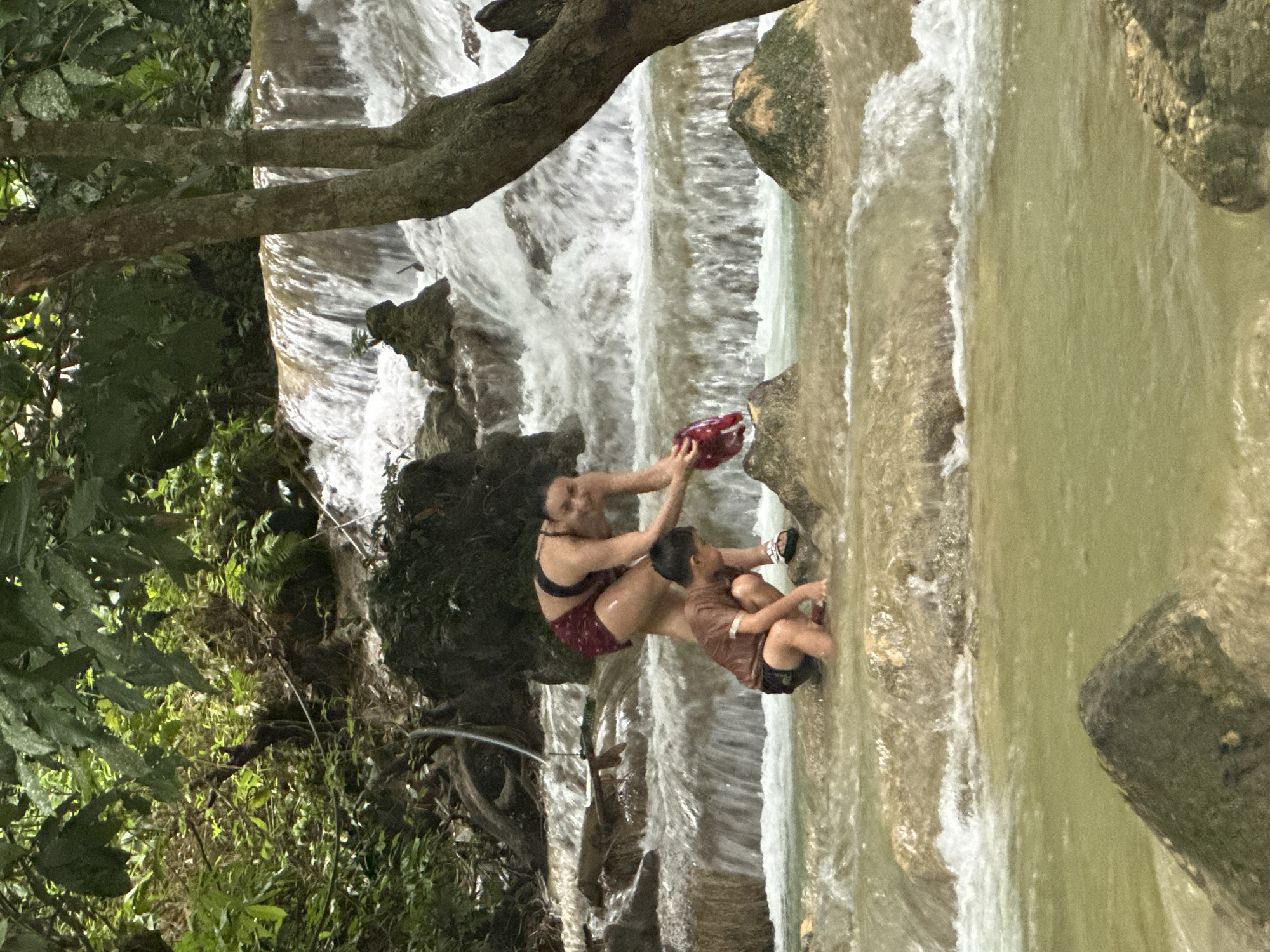 A woman and a young boy are sitting on rocks next to a waterfall. The woman is wearing a black sports bra and red shorts, and the boy is wearing a brown shirt and black shorts. The woman is holding a red object, possibly a bag, and the boy is reaching out towards it. They appear to be playing or interacting in the water.
The scene is a lush, natural environment with a cascading waterfall and surrounding greenery. The water is clear and flowing over the rocks, creating a refreshing atmosphere. The lighting suggests it is daytime, with sunlight filtering through the trees. The overall mood seems playful and relaxed, with the woman and child enjoying their time together in nature. The location context places this scene in Cổ Lũng Commune, Vietnam. There is no visible text in the image.