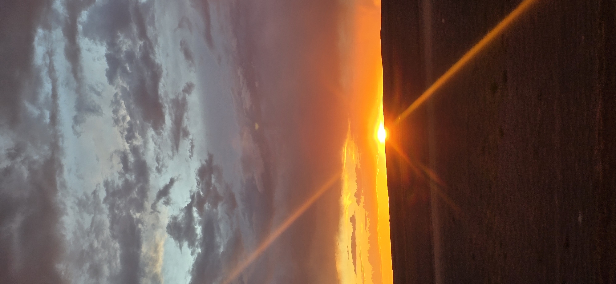 The image depicts a sunset over a desolate landscape, likely in or near the city of Tsogt-Ovoo, Mongolia. The sky is filled with a dramatic display of clouds. The upper portion of the sky is a cool, grayish-blue, with darker, textured clouds scattered throughout. As the eye moves down, the clouds transition to hues of pink and deep orange, indicating the setting sun.

The sun itself is visible as a bright, intense orb just above the horizon line, casting a brilliant orange glow. Rays of sunlight, appearing as sharp, diagonal beams of orange light, extend from the sun across the sky and down towards the foreground.

The foreground is characterized by a dark, undulating horizon line, suggesting rolling hills or a vast, open plain. The ground itself is rendered in deep shades of brown and purple, appearing somewhat featureless and barren, consistent with a steppe environment. There are no discernible people, animals, structures, or vehicles in the image. The overall impression is one of natural beauty and vastness, with the dramatic sunset dominating the scene.