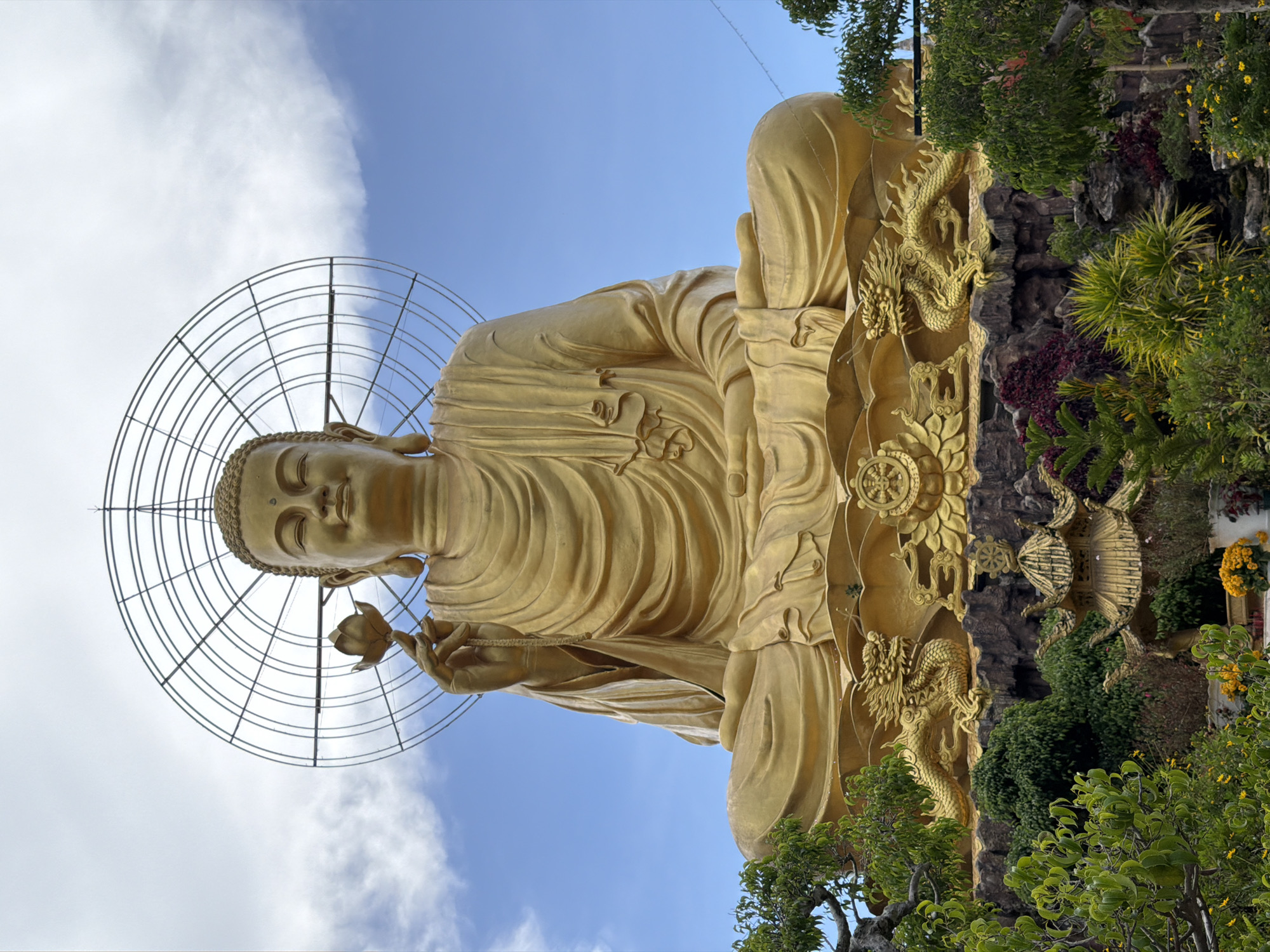 This is a full-body, eye-level, outdoor, vertical shot of a large, golden Buddha statue seated in a lotus position. The Buddha is holding a lotus flower in its right hand, which is raised. A circular, wire halo with radiating spokes is positioned behind the Buddha's head. The statue is situated on a rocky, tiered base adorned with golden dragon sculptures and intricate carvings. In the foreground, lush green foliage, colorful flowers, and a small, ornate golden pagoda are visible. The background consists of a bright blue sky with scattered white clouds. The scene is illuminated by natural daylight. The overall impression is one of grandeur and serenity, characteristic of a religious monument. The statue is located in Lâm Viên - Đà Lạt, Vietnam.