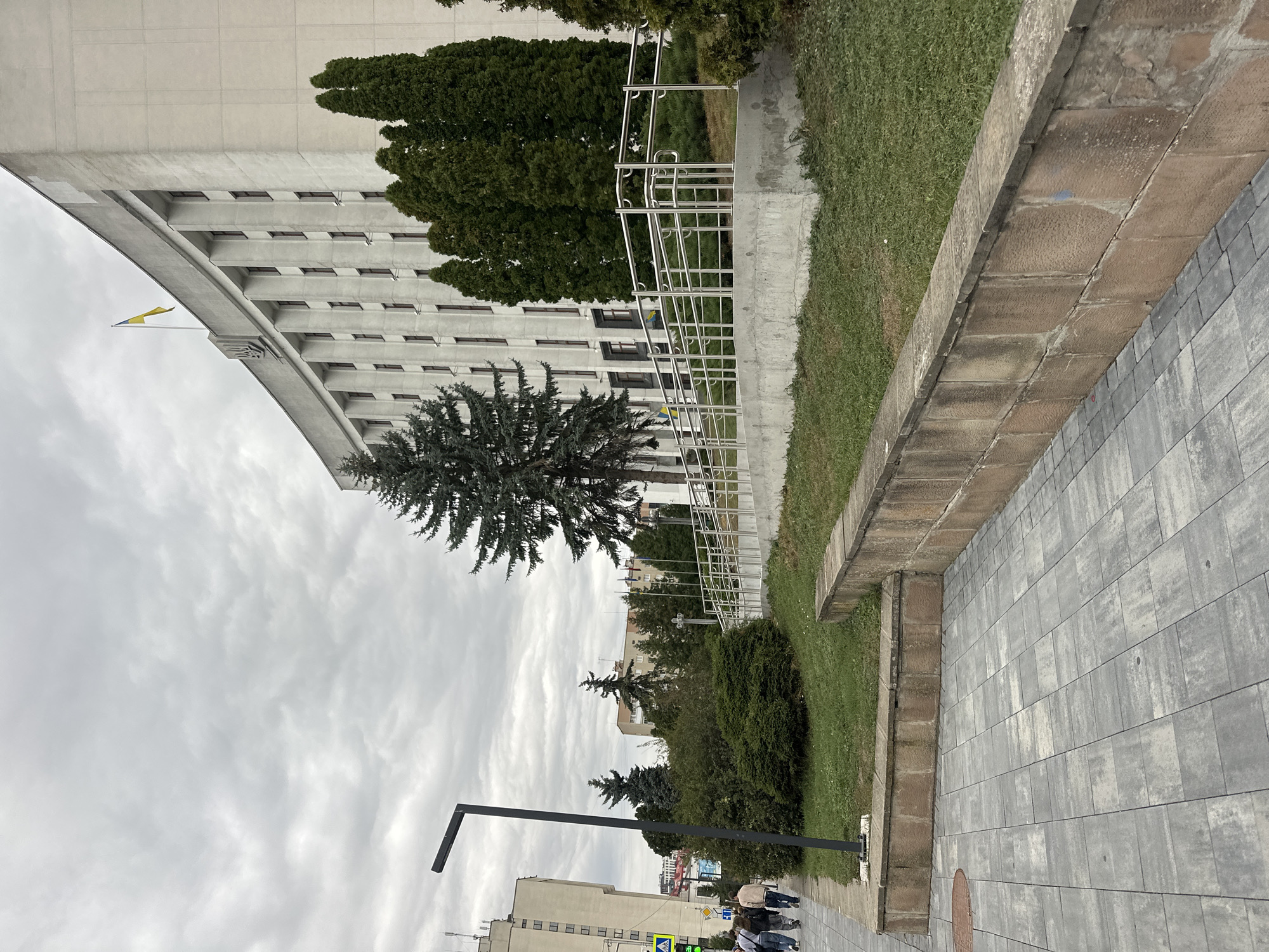 The image shows the Ternopil Regional State Administration building, located in Ternopil, Ukraine. The building is a large, curved, white structure with numerous windows and pillars. A Ukrainian flag flies from the top of the building. In front of the building, there is a sidewalk with grey paving stones, a grassy area, and a low stone wall. Several evergreen trees and bushes are planted in front of the building. In the background, other buildings and flags can be seen. A group of people are walking on the sidewalk. The sky is cloudy.