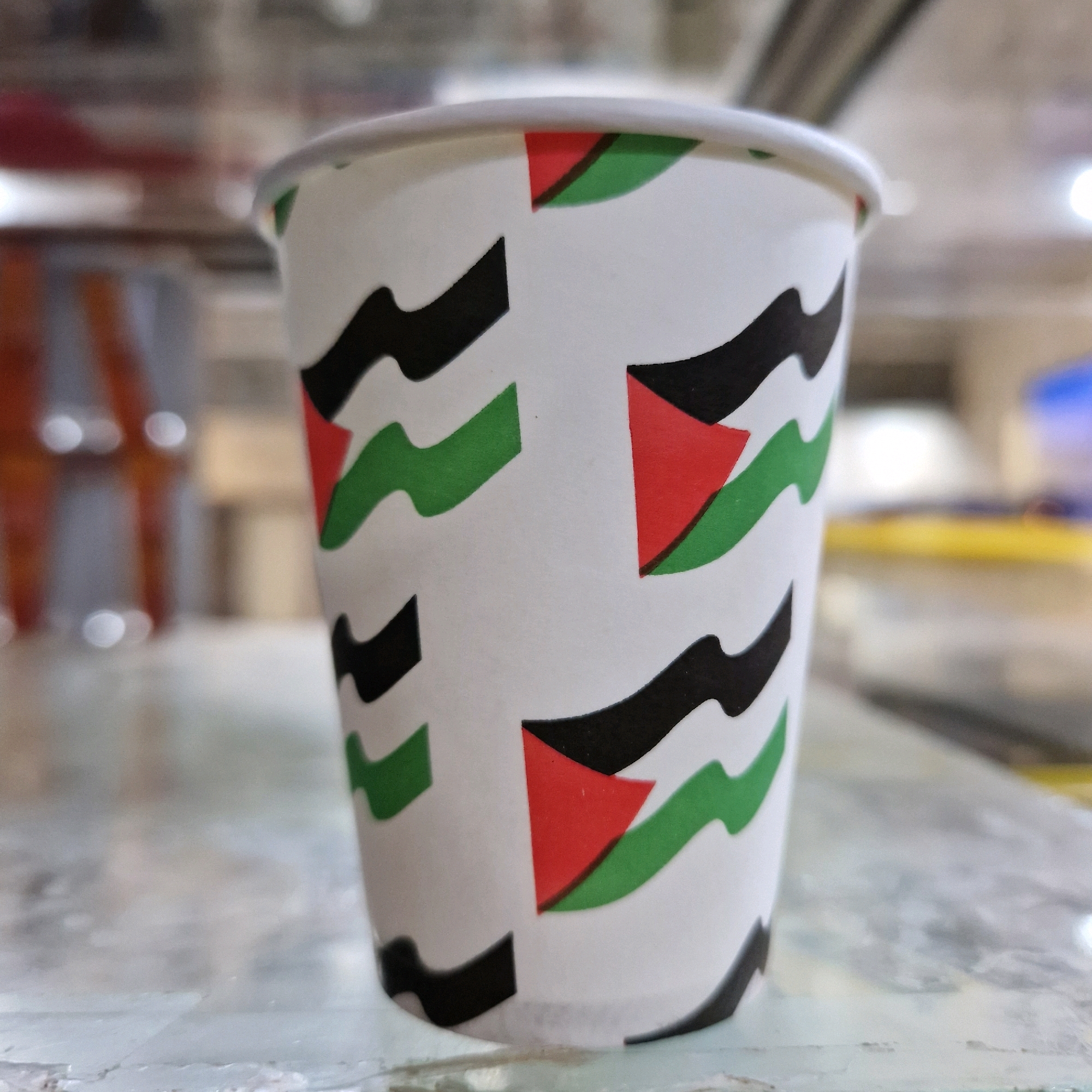 A close-up, shallow depth-of-field shot focuses on a white disposable cup decorated with a pattern that mimics the Palestinian flag. The pattern consists of horizontal black, white, and green stripes interspersed with red triangles. The cup is positioned on a speckled countertop, likely in an indoor setting. The background is blurred, showing hints of reflective surfaces and lights, suggestive of an interior environment such as a shop or cafe, possibly in the city of Makman Ben Ammar, Algeria. No people are visible.