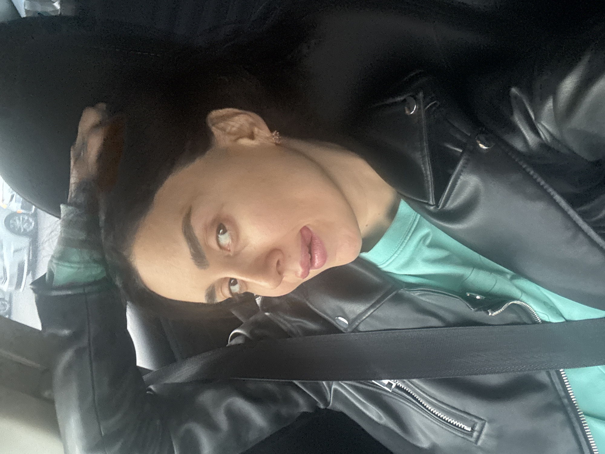 A young adult female is depicted in a close-up, slightly tilted portrait, appearing to be inside a vehicle. She has dark hair and dark, well-groomed eyebrows. Her gaze is directed upwards and to her left, with a pensive or slightly playful expression, her lips slightly parted to reveal a hint of her tongue. She is wearing a black leather-style jacket over a light teal or mint green t-shirt. A black seatbelt is prominently visible, crossing diagonally from her left shoulder down towards her right hip. The background is dark and out of focus, indicating the interior of a car, with a blurry glimpse of what appears to be a car window and the external environment, possibly other vehicles, on the left side of the frame. The lighting suggests it is daytime. The location context for this scene is Tbilisi, Georgia. No visible text is present in the image.