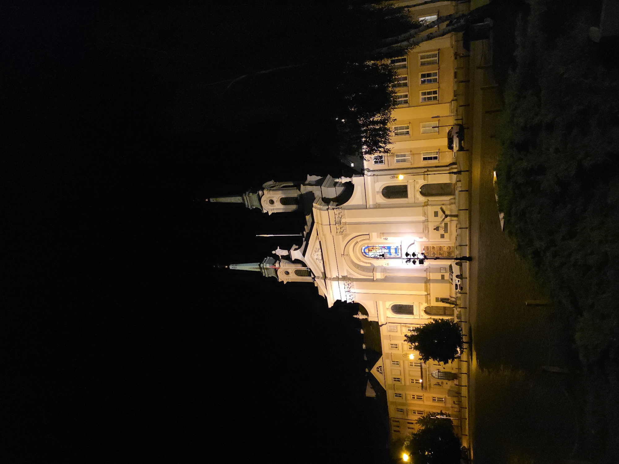 The image shows St. Anne's Church in Warsaw, Poland, at night. The church and adjacent building are illuminated. A street light provides additional lighting. There are trees and a vehicle near the building. The rest of the image is dark.