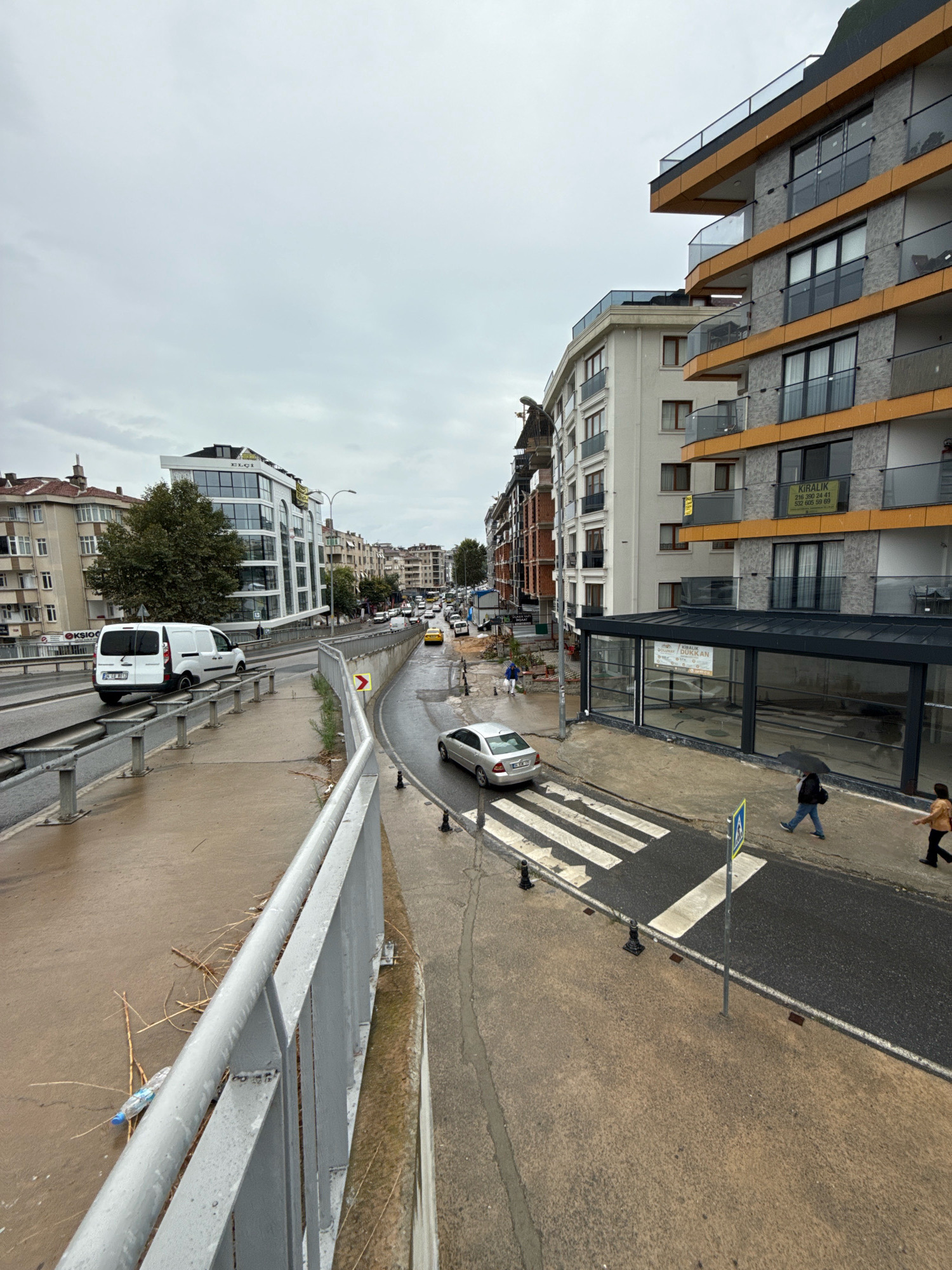 The image captures a city street scene on an overcast day, with buildings lining both sides of a road. In the foreground, a gray metal railing runs along a raised walkway or overpass, with debris and a plastic bottle scattered on the wet ground below. A white van is visible on the road to the left, and further down the road, a silver car is stopped at a pedestrian crossing. Several other cars are seen in traffic further down the street. On the right side of the road, a modern multi-story building with orange accents and balconies stands next to a row of older buildings. People are walking on the sidewalk, one carrying an umbrella. The overall environment appears to be a moderately busy urban area.