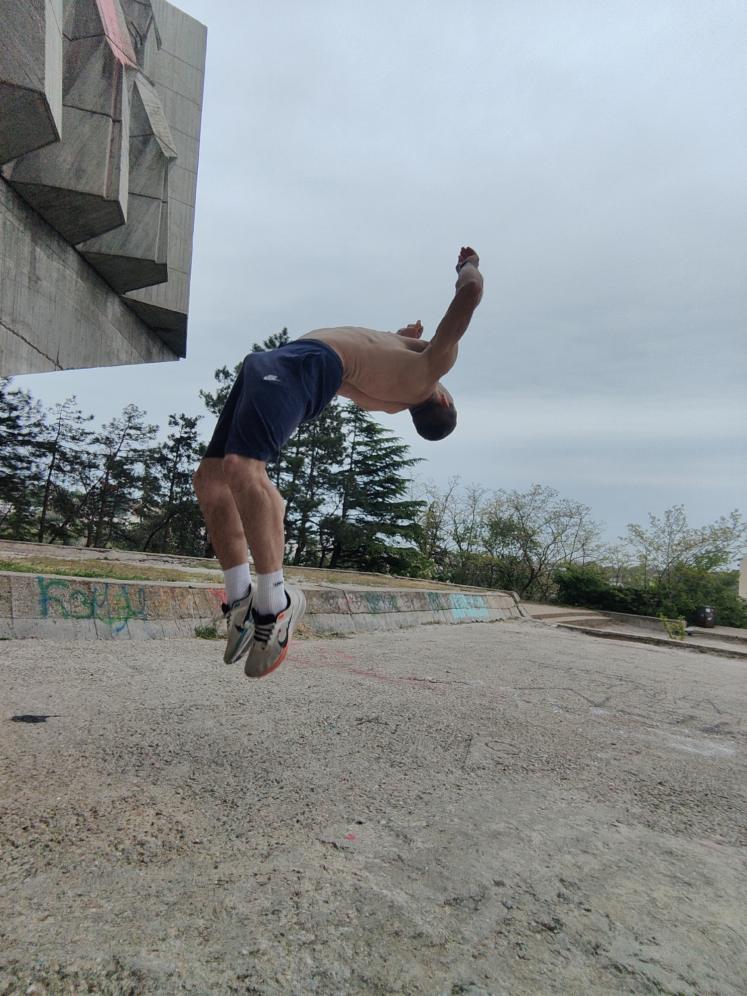 A shirtless man in navy shorts is performing a backflip in front of a concrete monument, located in Varna, Bulgaria. The monument is partly visible in the upper-left corner of the image and appears to have a relief or sculptural design. The surrounding environment includes trees, a low concrete wall with graffiti, and a concrete surface. The sky is overcast. The man is wearing white socks and athletic shoes, and his arms are raised above his head in mid-flip.