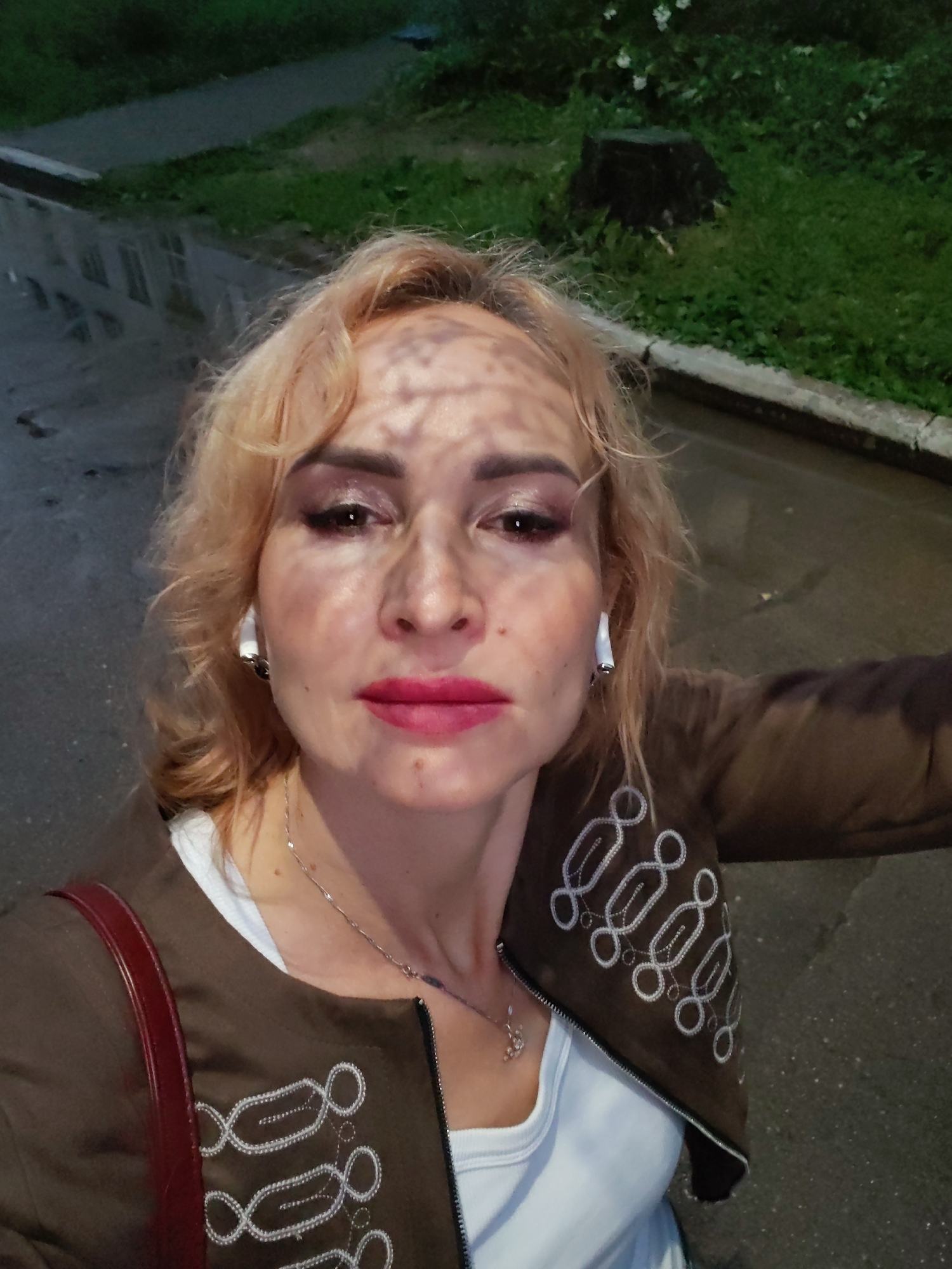 In Chaikovsky, Russia, a blonde woman with bright pink lipstick and visible eyeshadow stands close to the camera, facing forward. She wears a white top underneath a brown jacket adorned with white decorative stitching. A red purse strap is visible over her left shoulder. The background shows a wet, paved area reflecting streetlights, suggesting it might be nighttime or dusk, with a grassy area and a tree stump to the right. The woman has white earbuds in both ears.