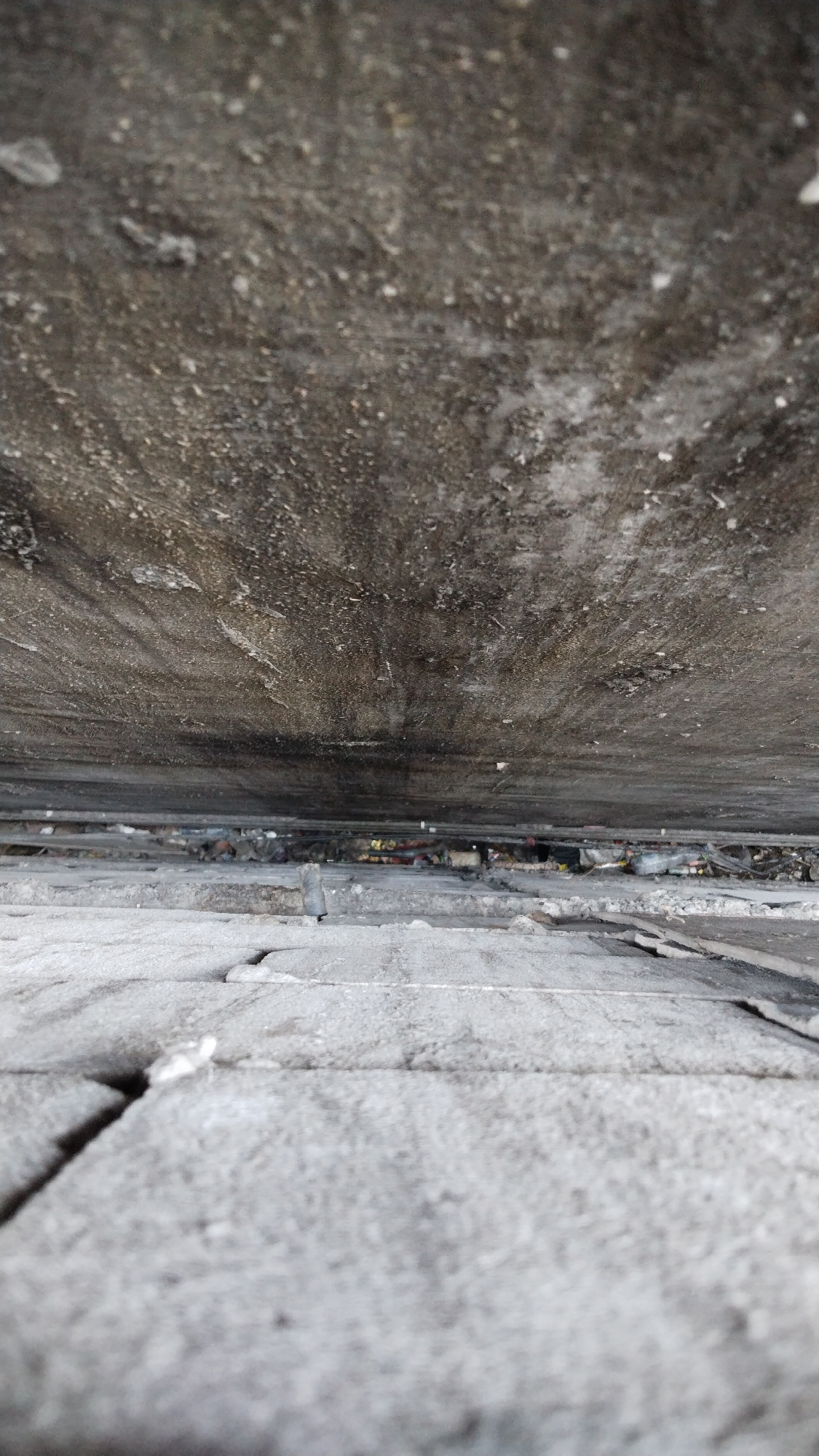 The image shows a perspective looking down from a height onto a surface of gray concrete or stone blocks. The blocks are roughly rectangular and appear to be laid in a pattern with visible grout lines. The surface in the foreground is more in focus, with cracks and texture visible in the blocks. As the perspective recedes into the distance, the surface becomes less distinct.
Above this surface, there is a dark, rough, and textured area, possibly a wall or an overhang, which also recedes into the distance. Between the foreground surface and the overhead surface, there is a gap where debris and what appear to be colorful scraps of material are visible. This suggests a narrow space, perhaps an alleyway or a gap between structures.
The scene appears to be an outdoor urban environment. The lighting suggests it is daytime, but the quality of light is not particularly bright, given the dark tones of the overhead surface and the general muted colors.
The image does not contain any people or clear indications of specific actions or interactions. It is a static view of an urban architectural detail and its immediate surroundings.
The context provided places this scene in Banjarsari, Indonesia, which is a subdistrict within Surakarta (Solo), Central Java. This suggests that the architectural style and materials, such as the concrete or stone blocks and the rough texture of the overhead surface, are consistent with typical urban infrastructure found in this region of Indonesia.