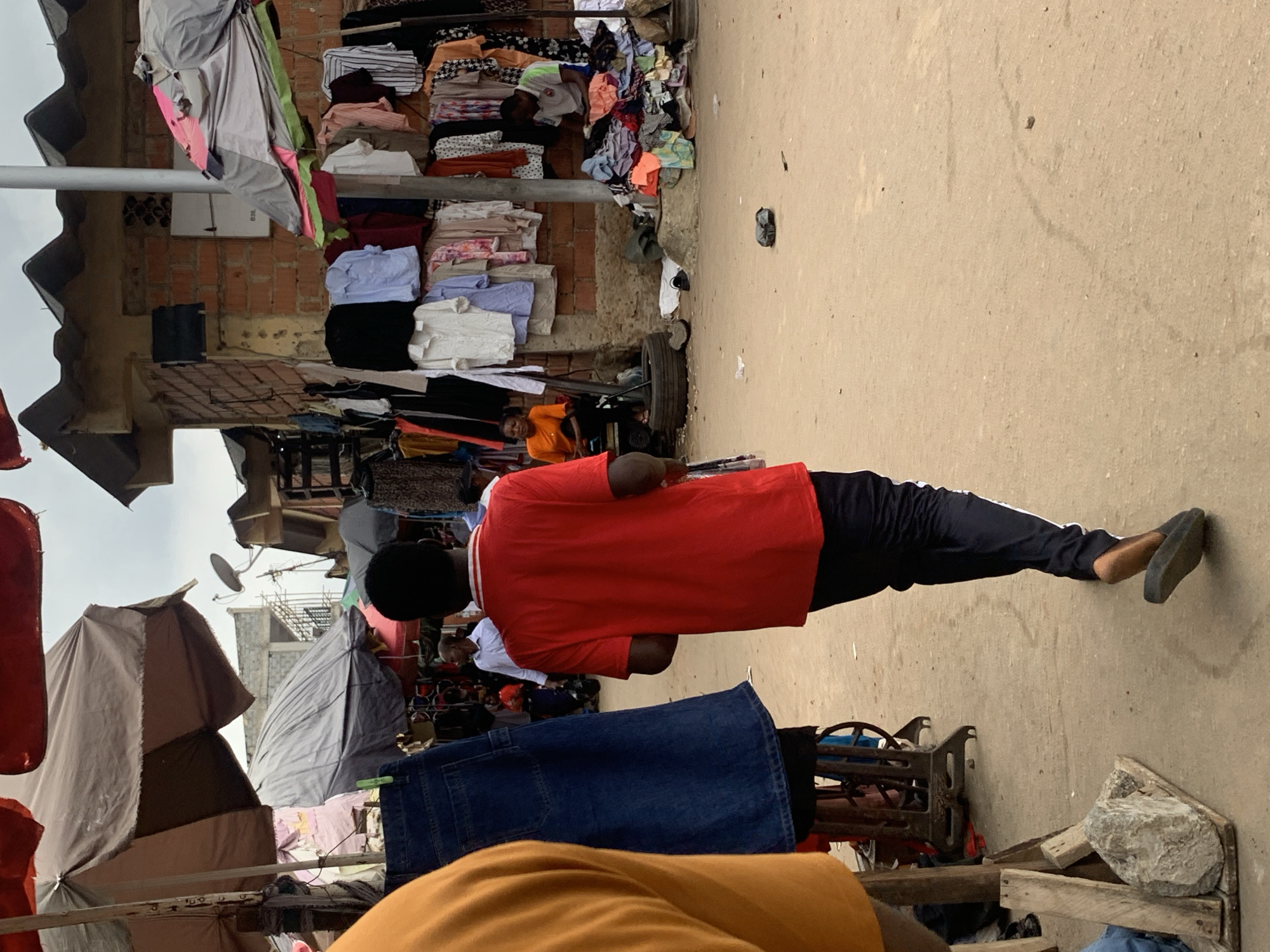 This image captures a bustling street scene in Abuja, Nigeria, likely a marketplace. The primary subjects are several people, including a person in the foreground wearing a red t-shirt and dark pants, facing away from the camera. Another individual in an orange shirt is visible further back. The scene is filled with a variety of objects, predominantly clothing. Piles of folded shirts and other garments are displayed on shelves and draped over structures. Several awnings and tarps provide shade, suggesting an outdoor market environment. In the background, hints of a brick building and some satellite dishes indicate a more urban setting.
The activity depicted seems to be the operation of a market, with goods displayed for sale and people presumably browsing or working. The time of day appears to be daytime, given the ambient light, and the weather seems overcast, as there are no strong shadows. The presence of clothing racks, folded fabrics, and what looks like a sewing machine part suggests a place where clothing is sold or possibly manufactured. The overall impression is one of a lively and functional commercial space. No visible text is discernible in the image.