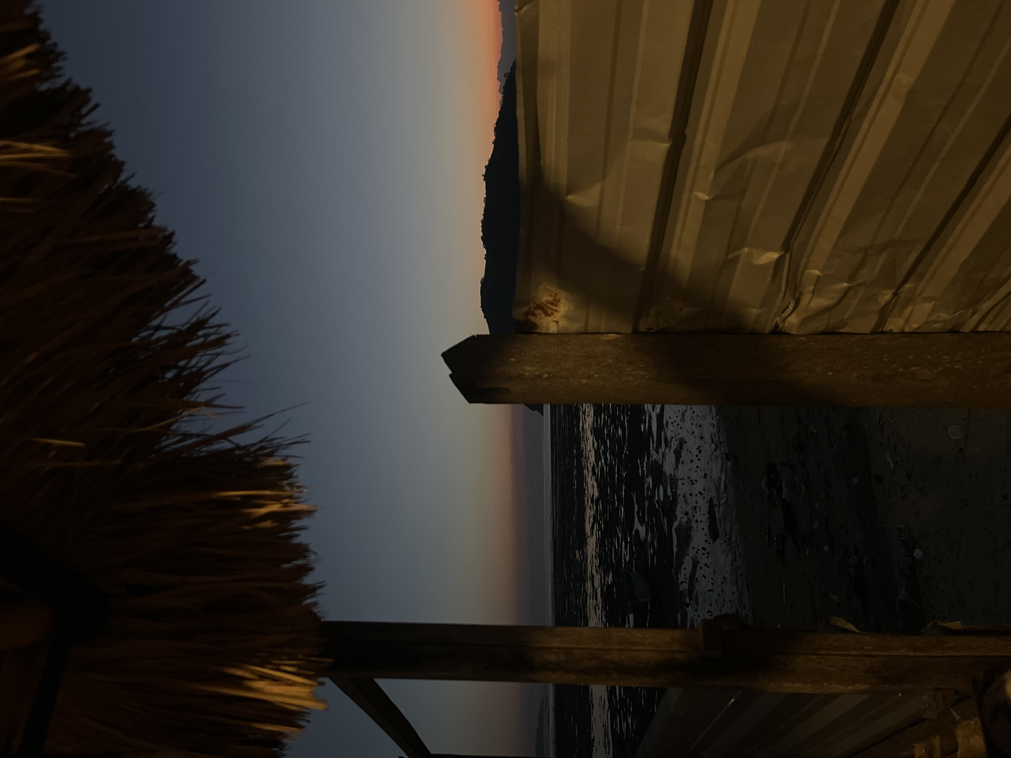 The image depicts a twilight scene on the beach in Kertasari, Indonesia, viewed from under a thatched roof structure. In the foreground, a wooden beam and corrugated metal wall partially obscure the view of the beach. The beach itself is partially submerged, with shallow water reflecting the dusky sky. A small boat is visible in the water. In the distance, a silhouette of a hilly landscape is outlined against the horizon, where the sky transitions from a deep blue to hues of orange and pink. The overall atmosphere is serene, characteristic of a coastal sunset.