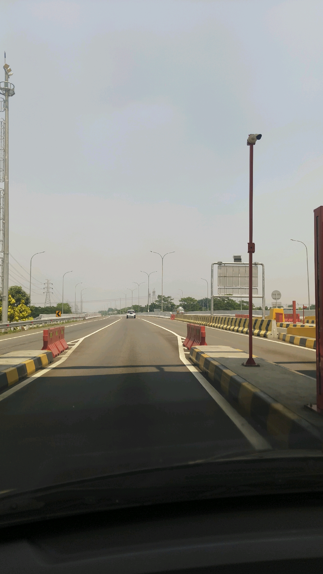 The image depicts a view from the driver's seat of a vehicle on a multi-lane highway leading towards a toll plaza. The highway is paved and has clear white lane markings, with yellow and black striped barriers lining the edges. Red plastic traffic barriers are strategically placed. Streetlights with high-intensity discharge (HID) lamps are visible along the road. In the distance, a white car is driving on the highway, and a large electronic sign structure is positioned above the toll plaza. The sky is overcast. Power lines and transmission towers are visible in the background, suggesting an urban or semi-urban environment, consistent with the location of Situ Gadung, Indonesia. The scene is captured on a bright, possibly hazy, day.