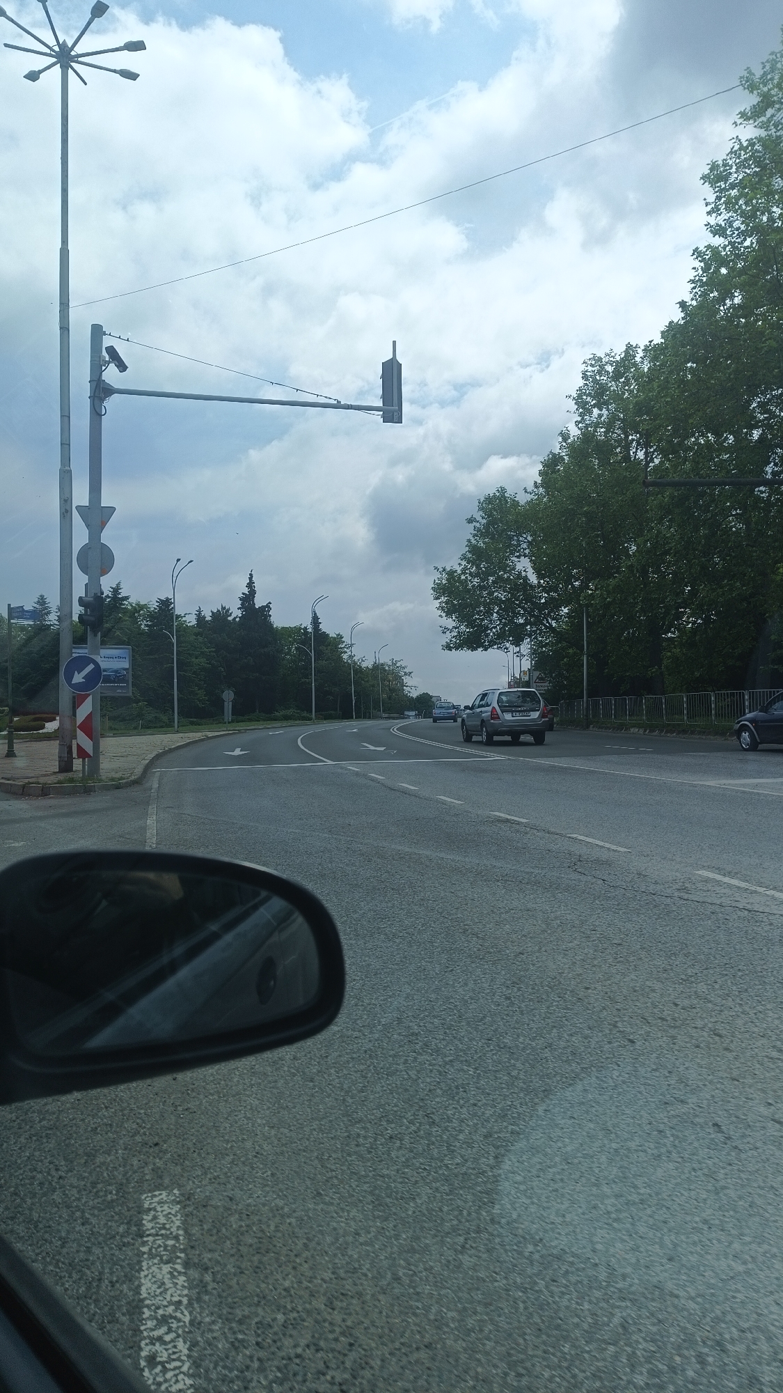 The image captures a street scene in Burgas, Bulgaria. A road curves to the right, marked with lane direction arrows. Several vehicles are present, including a silver SUV and a dark sedan. Roadside elements include streetlights, signage, trees, and a signal light suspended over the road. A rearview mirror of the vehicle from which the photo was taken is visible in the foreground. The sky is overcast with light and dark clouds.