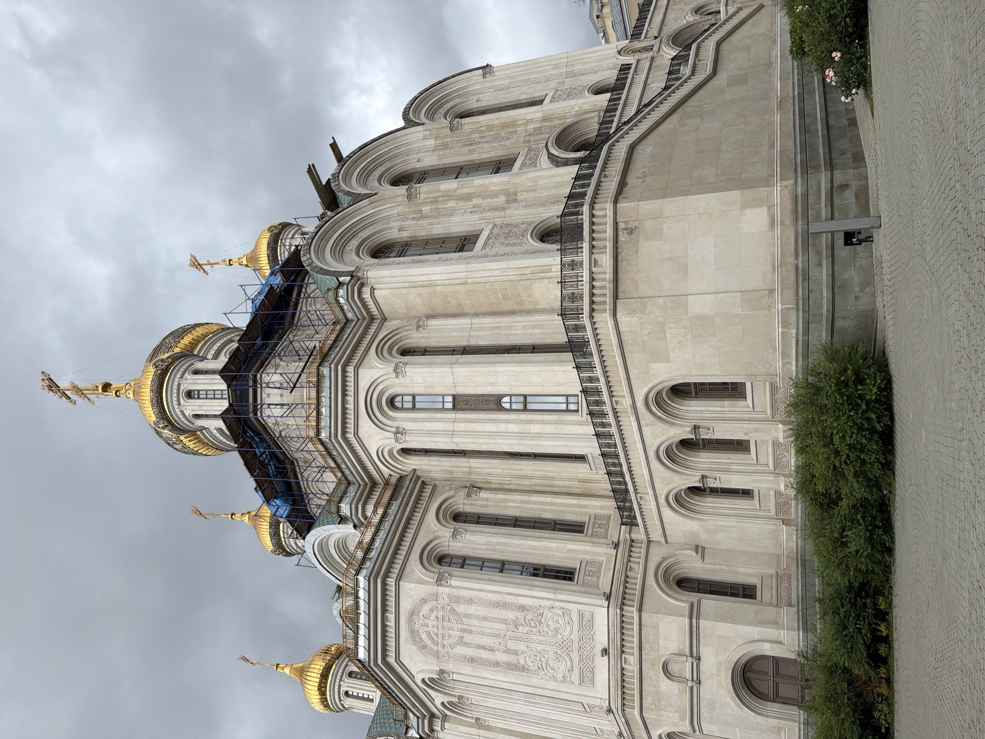 An exterior low-angle, medium shot captures the side of a large, ornate, light-colored stone church with golden domes and crosses. The sky is overcast and gray. Scaffolding is visible on the upper portion of the church, surrounding some of the domes. The church features arched windows and decorative carvings. A black metal railing lines a raised walkway or balcony. Below the church, green bushes and a paved area with a circular pattern are visible. Stairs with a railing lead up the side of the church. In the lower right, a small cluster of pink roses is in bloom. A figure is partially visible near a small structure at the base of the church.