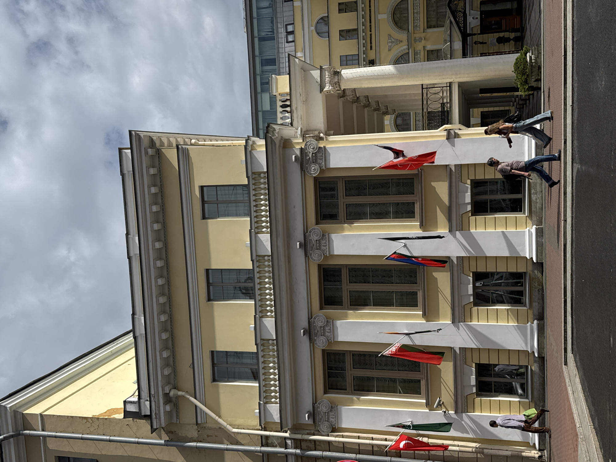 The image depicts a view of the building located at 17 Nevsky Prospekt in Saint Petersburg, Russia. The building is a pale yellow, multi-story structure with classical architectural elements, including columns and decorative molding. Several flags are displayed on poles attached to the building's facade. In the foreground, a man in a purple shirt and brown pants walks to the left, carrying a green bag. Further to the right, a man and a woman walk in the same direction. The man wears a brown t-shirt and blue jeans, and the woman wears a white top and blue jeans. They are walking on a red brick sidewalk adjacent to the building. The sky above is cloudy.