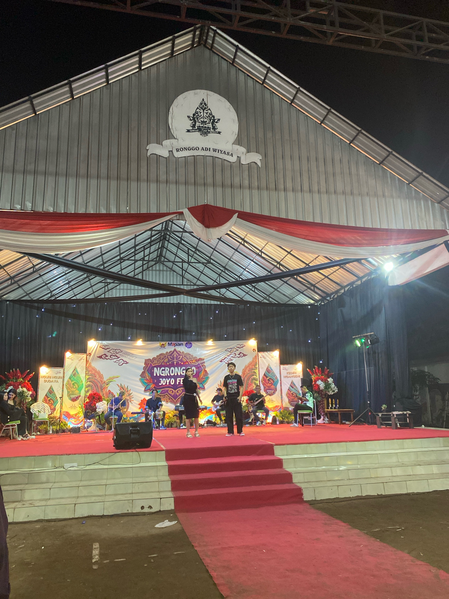 The image depicts an outdoor stage setup at night, likely for a cultural event or performance in Kediri City, Indonesia. A large, corrugated metal structure with a triangular roof forms the backdrop, adorned with a circular logo featuring traditional Javanese motifs and the text "RONGGO ADI WIYASA". Red and white fabric drapes are suspended from the roofline. The stage itself is elevated and covered with a red carpet, with a set of red carpeted stairs leading up to it.
On the stage, a banner with colorful graphics and the text "NGRONGGO JOYO FE" is prominently displayed. Several individuals are present on the stage, some performing or playing musical instruments, while others appear to be audience members or performers observing. A woman in a black dress is center stage, seemingly addressing the audience or performing. To her left, a man in a black t-shirt and pants stands. A group of people are seated in the background, some playing instruments like guitars and drums. To the left of the stage, a speaker unit is visible.
The surrounding environment appears to be an open area, possibly a plaza or field, with the ground covered in dirt and some scattered debris. Lighting from stage lights and surrounding illumination creates a dramatic atmosphere. The overall impression is of a vibrant cultural gathering in an urban setting.