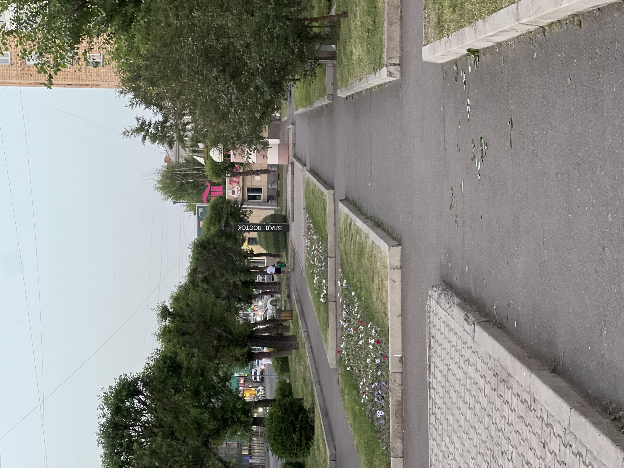 This is a photograph of a pedestrian walkway in Krasnoyarsk, Russia. The walkway is lined with grass and flower beds, and mature trees provide shade. A paved road runs alongside the walkway, and a brick building is visible in the background. The sky is overcast, suggesting a cloudy day. There is a black sign with white Cyrillic text that reads "ВЛАД ВОСТОК", which translates to "Vladivostok". This is likely a street name or a directional sign. In the distance, there are hints of traffic and some commercial establishments. The scene appears calm and suggests a typical urban environment. The time of day is unclear due to the overcast sky, but it seems to be daytime. No specific event or activity is clearly depicted, other than people potentially walking or sitting in the area.