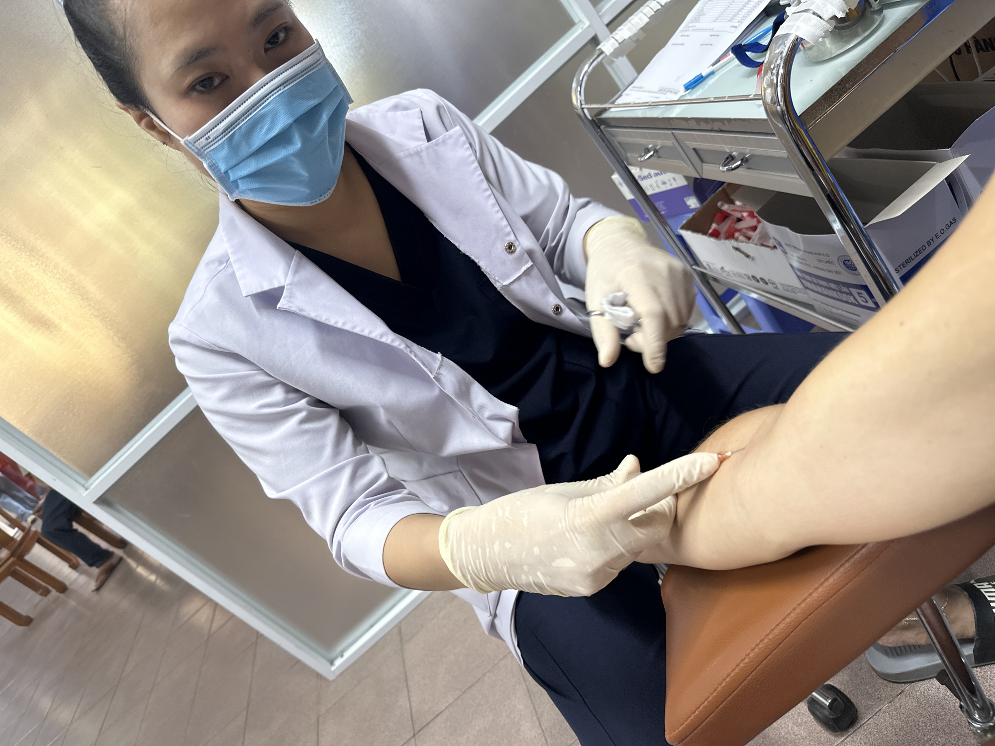 A person in a white lab coat, blue face mask, and beige gloves is preparing to inject a needle into the arm of another person seated on a brown chair. The scene appears to be a medical setting, possibly a clinic or hospital, with medical equipment visible in the background, including a metal cart with various supplies. The floor is tiled, and there are frosted glass partitions and wooden chairs visible. The overall impression is one of a routine medical procedure taking place in an indoor, sterile environment.