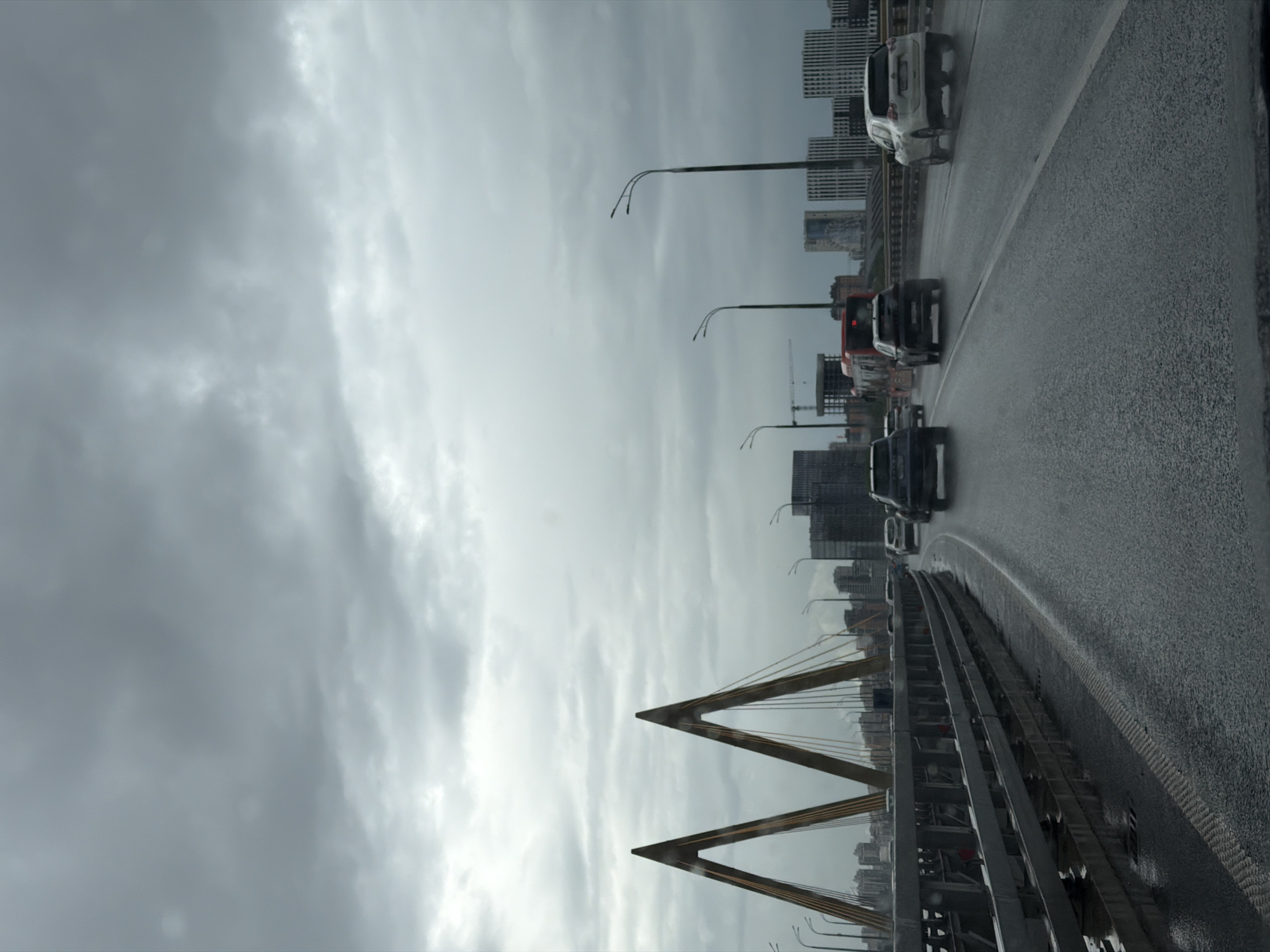 The image depicts a view from a vehicle traveling on the Mileniy Bridge in Kazan, Russia, on a cloudy day. The bridge's A-shaped pylons are visible, as are city buildings in the background. Cars are moving in the same direction on the wet road. Street lamps line the bridge.
