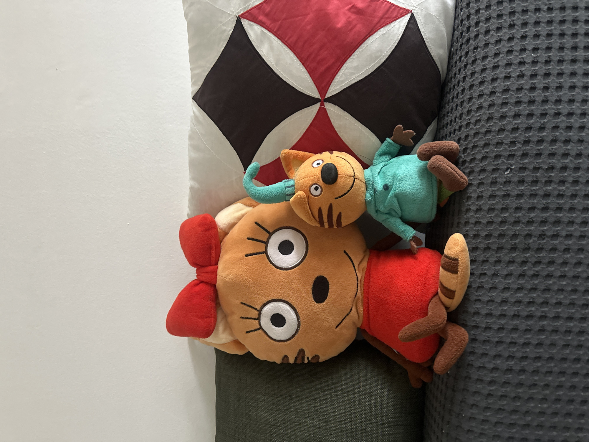 The image displays two plush toy figures of anthropomorphic cats seated on an upholstered surface against a plain white wall. The larger plush figure, positioned on the left, is orange-brown, wears a red dress, and has a red bow on its head. Its facial features include large white eyes with black pupils and black eyelashes, a black nose, and a thin black smile, along with brown stripes on its cheeks. Its tail is striped orange-brown and dark brown. The smaller plush figure, on the right, is also orange-brown, wears a teal long-sleeved shirt and green pants, and has a teal, bent, antenna-like appendage on its head. Its face features similar white eyes with black pupils, a black nose, a wider black smile, and brown cheek stripes. Both plush toys are situated on a dark gray, waffle-textured fabric surface, which is likely a sofa or bed covering. Behind the larger plush figure is a dark green solid-colored cushion. Behind the smaller plush figure is a white square cushion featuring a patterned design with four quadrants, two black and two red, bordered by white quilting. The exact location within the city of Budva, Montenegro, is not discernible from the provided image, as the scene depicts an interior setting without identifying external features or landmarks.