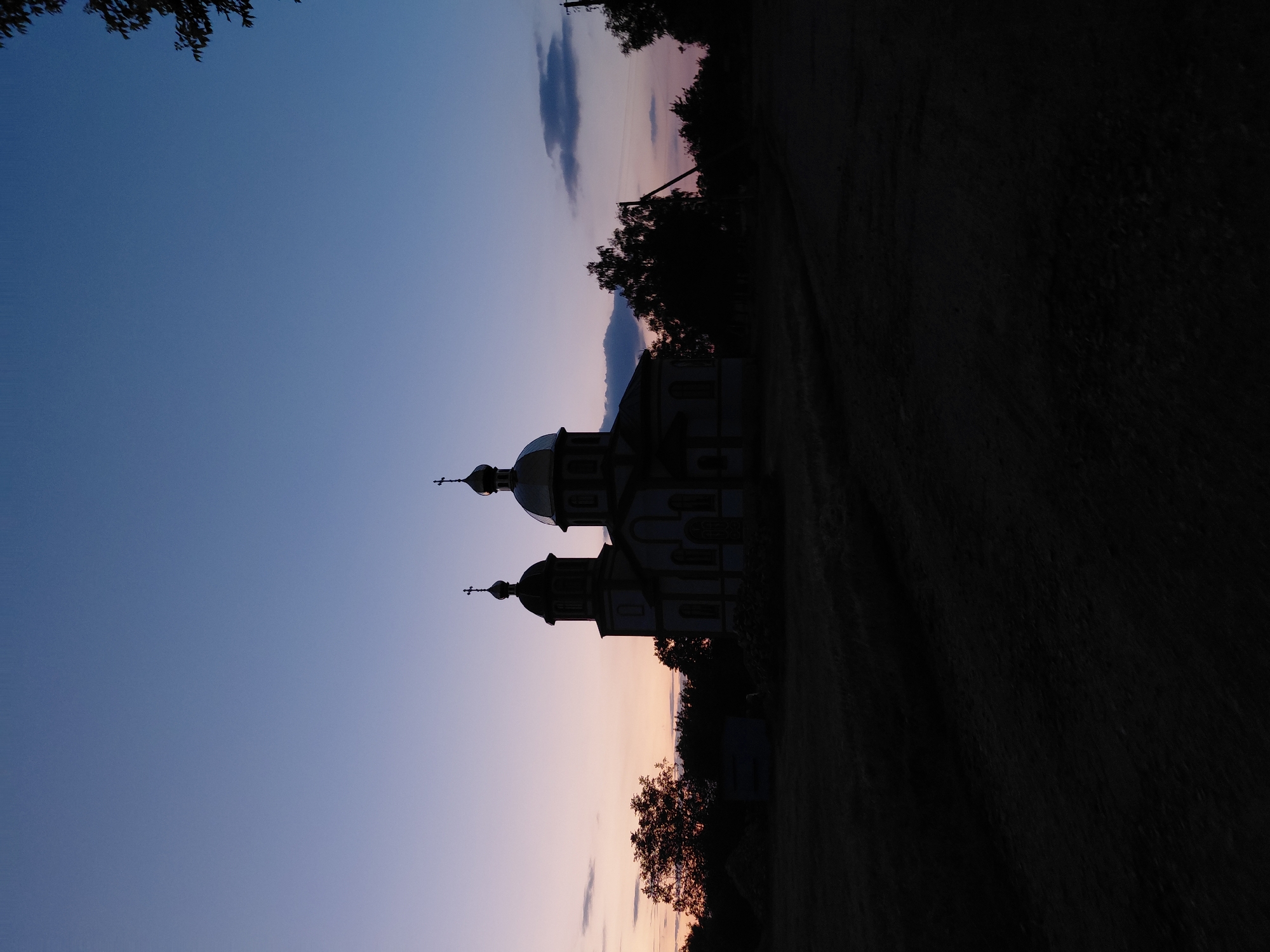 The image captures a silhouetted church in Ustyvytsia, Ukraine, during twilight. The sky transitions from a pale blue at the top to soft hues of pink and purple near the horizon, with a few scattered clouds. The church, with its characteristic domes and crosses, is positioned centrally, flanked by trees and vegetation that are also cast in shadow. The foreground consists of dark, textured ground, possibly a field or unpaved area, with some indistinct shadows indicating natural features or possibly haystacks. The overall atmosphere is serene and highlights the architectural form of the church against the fading light of dusk. There are no people visible in the image.