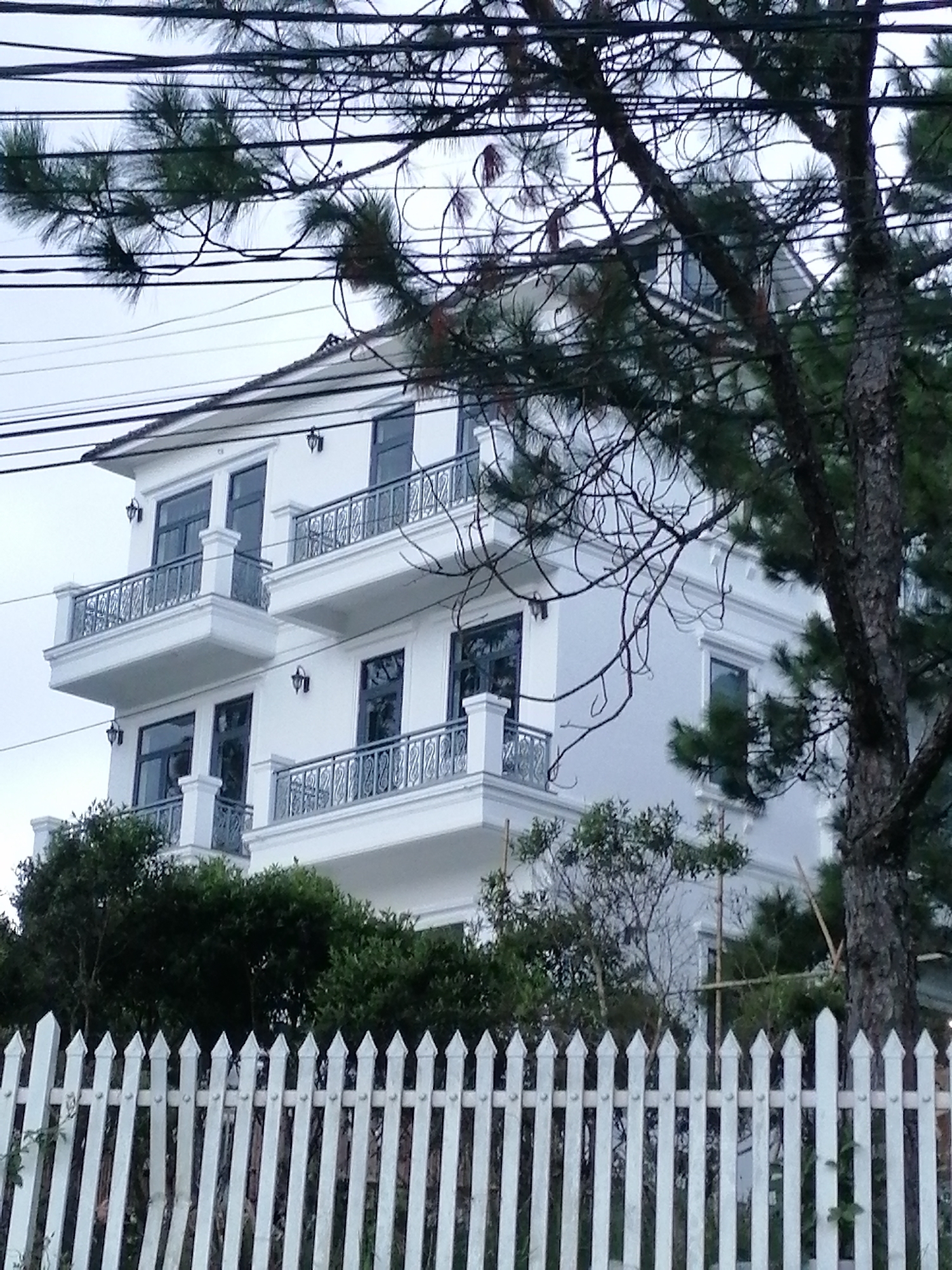 A white, multi-story house with balconies is partially obscured by pine trees and overhead power lines. The house has a pitched roof and balconies with ornate metal railings on each level. The windows are dark. A white picket fence runs along the bottom of the frame. The setting appears to be a residential area, possibly in a hilly or mountainous region given the pine trees. The sky is overcast. This scene is located in Xuân Trường - Đà Lạt, Vietnam.