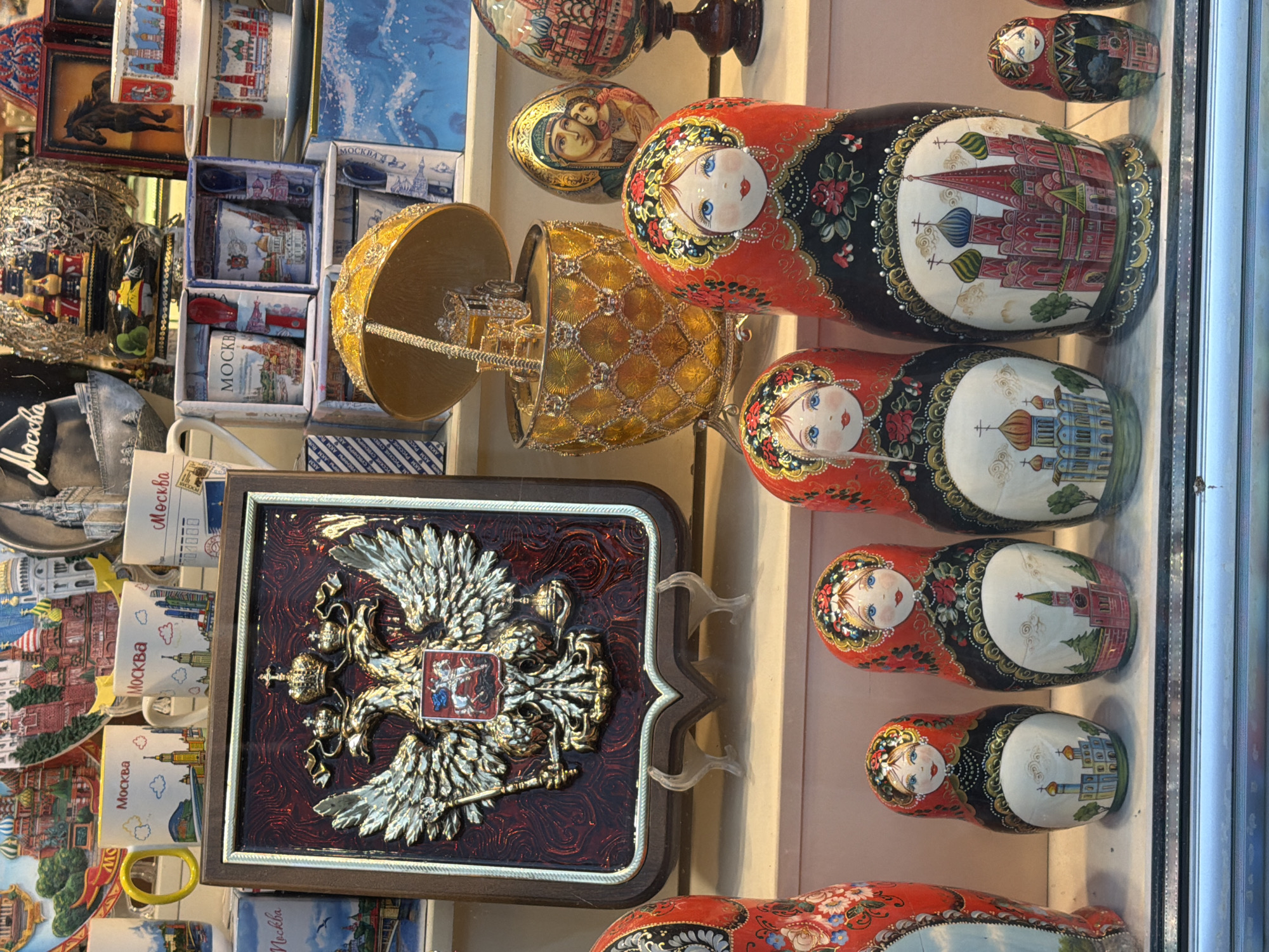 A close-up, eye-level shot captures a display of Russian nesting dolls and other souvenirs. The dolls, painted in vibrant reds and blues, feature traditional designs of women's faces and Russian architectural landmarks like cathedrals. They are arranged in a row on a white surface, with larger dolls in the foreground and smaller ones towards the back. To the left of the dolls, a large, ornate wooden plaque with a golden double-headed eagle emblem is displayed on a stand. Behind the dolls, various other souvenirs are visible, including decorative eggs, mugs with cityscapes and Cyrillic script, and stacks of boxes. The overall setting suggests a souvenir shop or market stall.