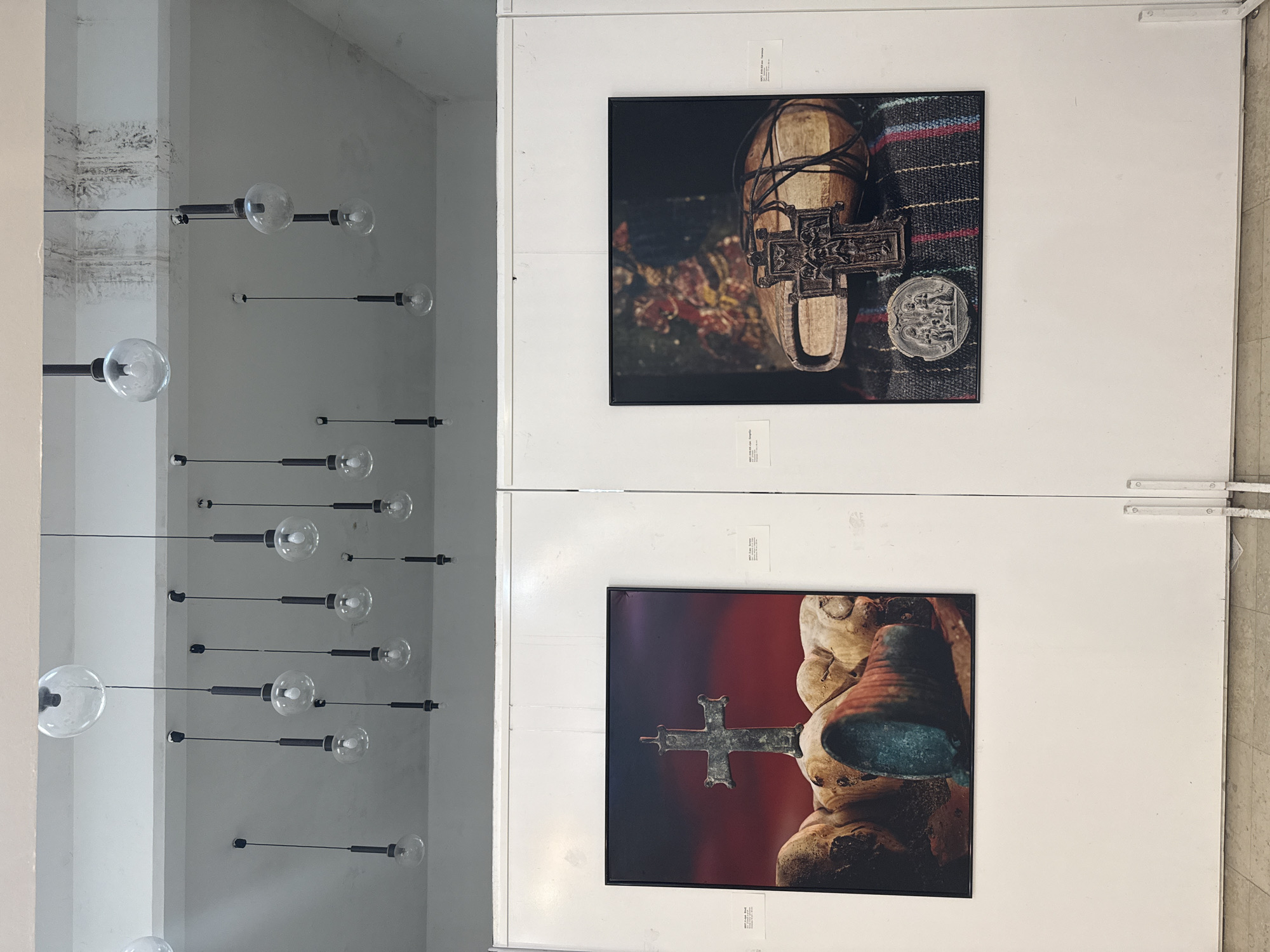 The image displays an indoor art exhibition space, likely a gallery in Bar, Montenegro. On the left, an array of modern, spherical glass light fixtures, each attached to a slender black rod, hang at varying lengths from a light-colored ceiling. The ceiling and a partial pillar on the far left show some textured, possibly aged or weathered, surfaces.
On the right side, two framed artworks are prominently displayed vertically on a pristine white wall.
The upper artwork is a photograph featuring a close-up of cultural artifacts. It shows a light-brown, boat-shaped or shoe-like wooden object wrapped with dark cords, resting on a woven textile with horizontal stripes of blue, red, and black. Positioned on the textile next to the wooden object are an intricately designed, dark metallic cross and a circular, carved metal medallion. The background is dark and softly blurred, with hints of red and green foliage.
The lower artwork is also a photograph, depicting a close-up of a weathered, organically shaped object, possibly carved wood or stone, with a patinated, green-tinged metallic cross resting against it or partially embedded within it. The background behind this composition is a soft, blurred red-orange hue.
Small white labels with black text are visible above and below each framed artwork. The upper label for the top artwork partially reads "2017 | 100x100 cm | Tsogou". The lower label for the bottom artwork also partially indicates the year "2017". The scene captures a contemplative atmosphere, characteristic of a cultural or artistic display.