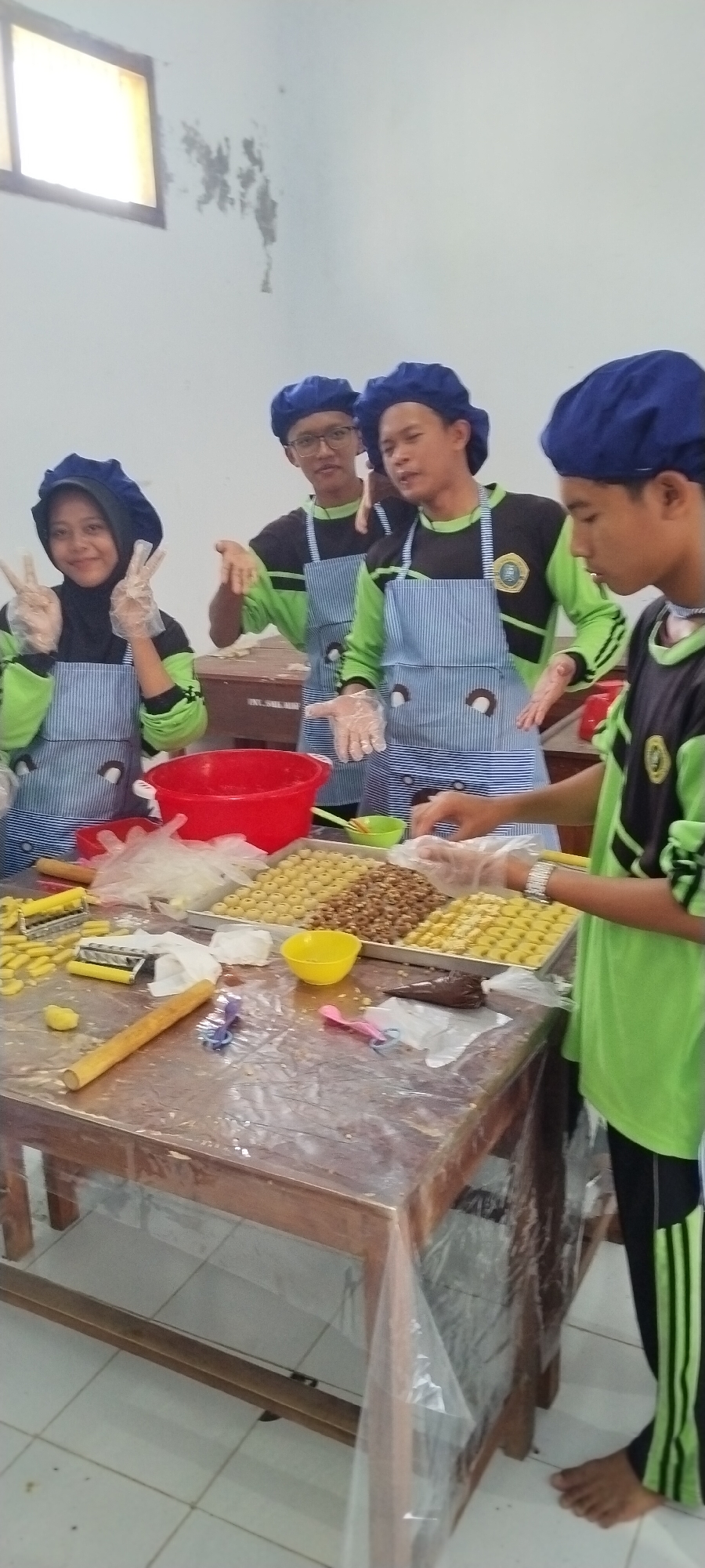 Four young individuals, two males and two females, are engaged in a culinary activity, possibly baking, at a wooden table covered with a transparent plastic sheet. They are all wearing blue hairnets and aprons over light green long-sleeved shirts. The individuals are in a brightly lit room with white walls and a window with diffused light.
In the foreground, the table is laden with ingredients and baking tools. There are trays filled with small, uniform, golden-brown items, possibly cookies or pastries, arranged in rows. Some of these items appear to be nuts. Other items on the table include a red bowl, small yellow bowls, plastic gloves, a wooden rolling pin, and what looks like a pastry bag filled with chocolate.
The individuals are actively participating in the process. One female on the left is smiling and making a peace sign with her fingers. A male in the center is looking towards the camera. Another male is looking down at the table, possibly working on the pastries. The fourth individual, a male on the right, is also focused on the task at hand.
The overall scene suggests a communal activity, likely a cooking or baking class or a recreational gathering focused on preparing food. The setting appears to be an indoor space with basic furniture and lighting, typical of a classroom or community hall. The location is identified as the city of Jombang, Indonesia.