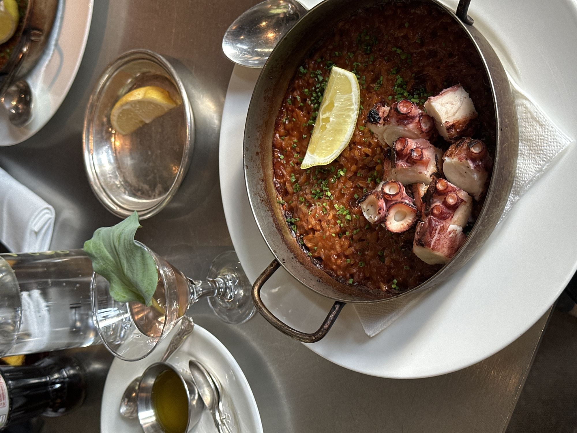 The image shows a table setting in a restaurant. A shallow metal pan containing a rice dish with octopus and a lemon wedge rests on a white plate with a white napkin. Next to the pan is a small silver dish with a lemon slice. A water bottle, a wine glass with a green leaf in it, a small silver cup with yellow liquid, and spoons are arranged on a white plate. Another white plate with food and a spoon is visible in the upper left corner. The setting is presumably a dining table in a restaurant, given the food presentation and utensils. There is no information about the specific location in London, United Kingdom in the image.