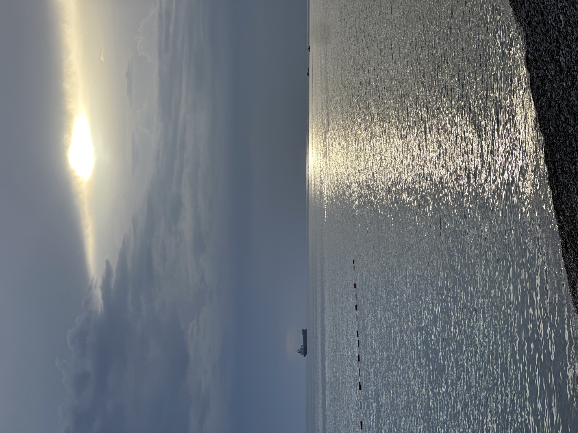 The image shows the Adriatic Sea at Šušanj, Montenegro. The sun is shining through clouds on the left side of the image. The water reflects the sunlight, with a ship visible in the distance. A line of red and white buoys is also visible in the water. The shoreline is visible on the right side of the image.