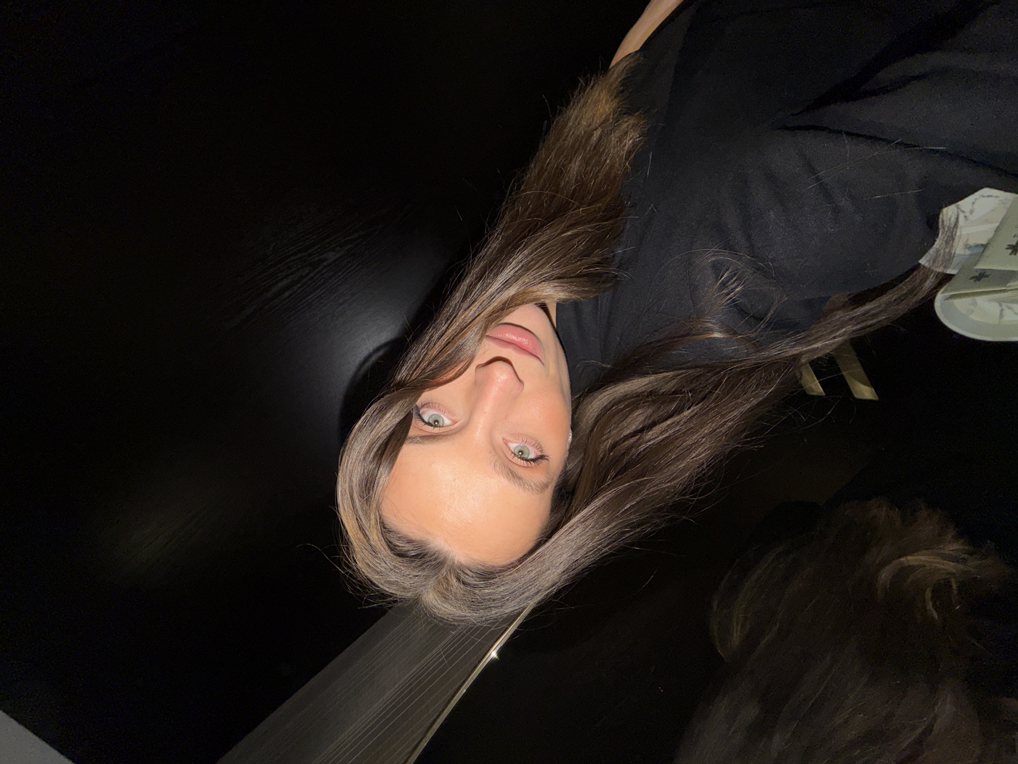 A woman with long brown hair and light-colored eyes is centered in the frame, looking directly at the camera. She is wearing a black shirt. The background is predominantly dark, suggesting an indoor setting with limited light. A wooden table is visible, also dark in color. In the lower right corner, there's a partial view of another person with brown hair. The location in Budva, Montenegro cannot be determined from this image.
