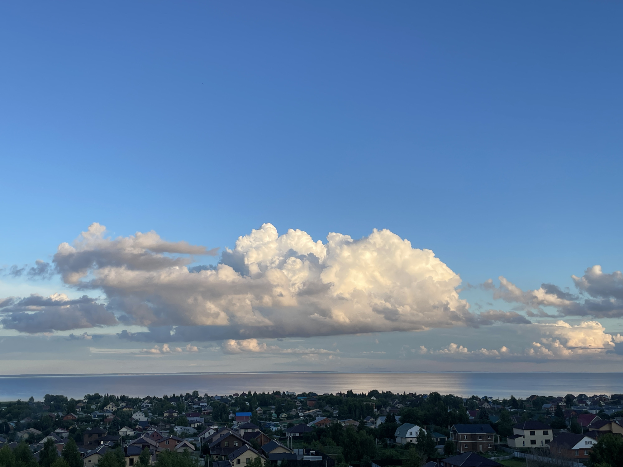 This is a high-angle, full shot of a cloudy sky over a coastal town. The sky is a vibrant blue, with a large, fluffy white cloud dominating the center of the frame. Below the sky is a body of water, likely a lake or sea, with a reflective surface. Along the shoreline, a town is nestled, with numerous houses of varying sizes and colors. Dense green trees surround the houses, creating a lush landscape. The houses appear to be modern, with pitched roofs and well-maintained yards. The overall impression is one of a peaceful, idyllic setting, with the grandeur of nature contrasted by the human settlement.