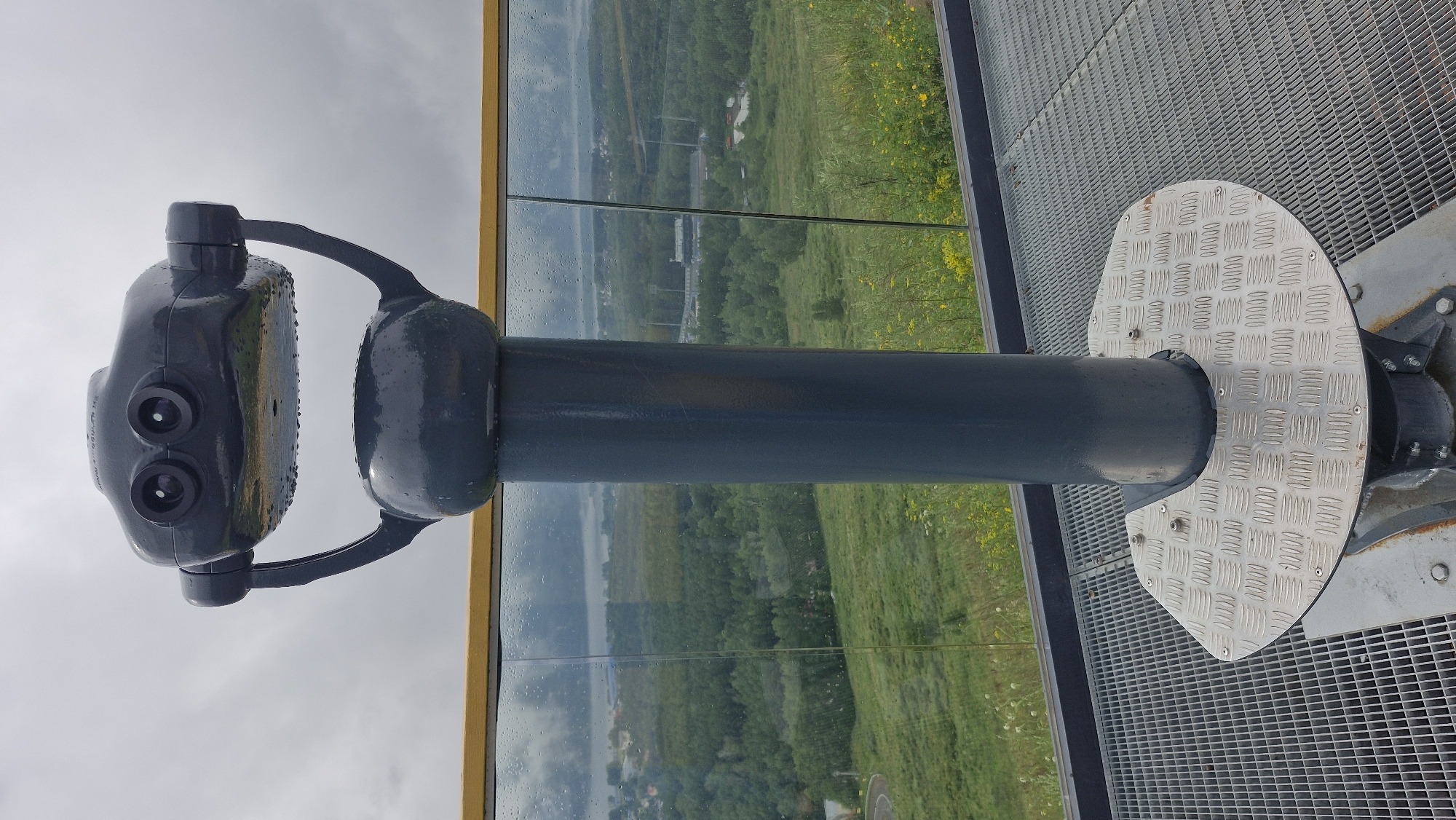 A dark gray, coin-operated binocular viewer stands on a metal grate platform, overlooking a landscape of green hills, trees, and a body of water under a cloudy sky. The binocular viewer is wet, with visible water droplets on its surface. A yellow-tinted glass railing separates the platform from the view. The platform has a diamond plate base where the binocular viewer is mounted. The background shows a rural or semi-rural area with some buildings and infrastructure visible in the distance. This binocular viewer is located at the Nikolayevskaya Sopka observation deck in Krasnoyarsk, Russia, a popular viewpoint offering panoramic views of the city and surrounding nature.