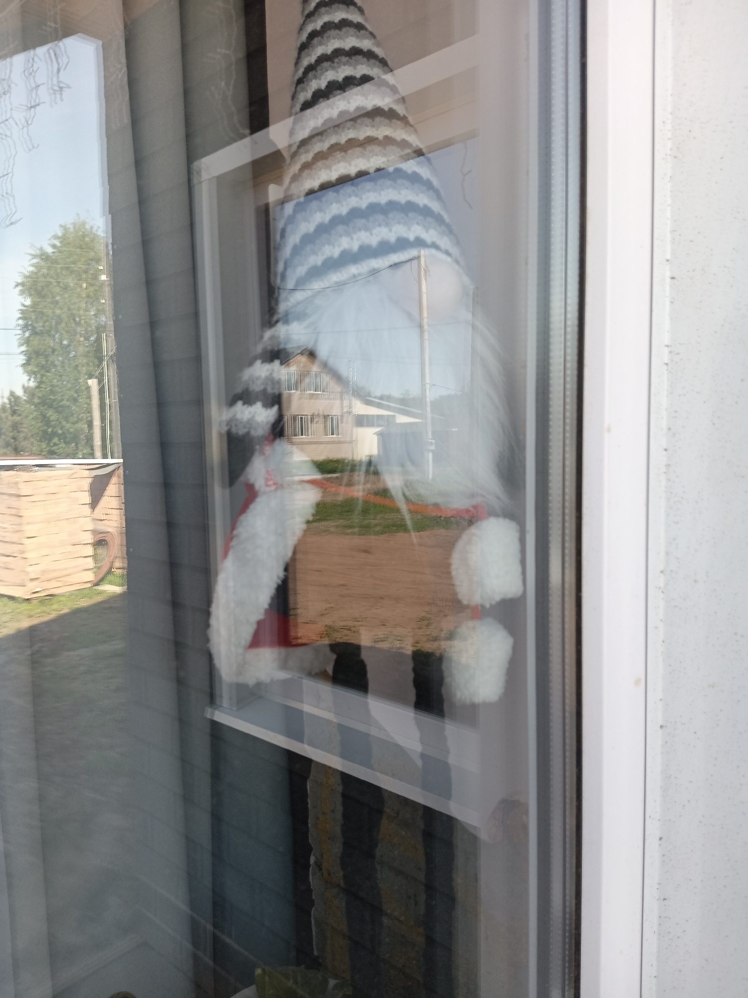 A gnome decoration hangs inside a window. The gnome has a tall, striped hat, a long white beard, and a red body with white trim. The window reflects the outside environment, including a building, trees, and a shed. The location appears to be Фоки, Russia, based on the metadata. The photo is taken from inside, looking out through the window. The curtains are visible on the left side of the window.