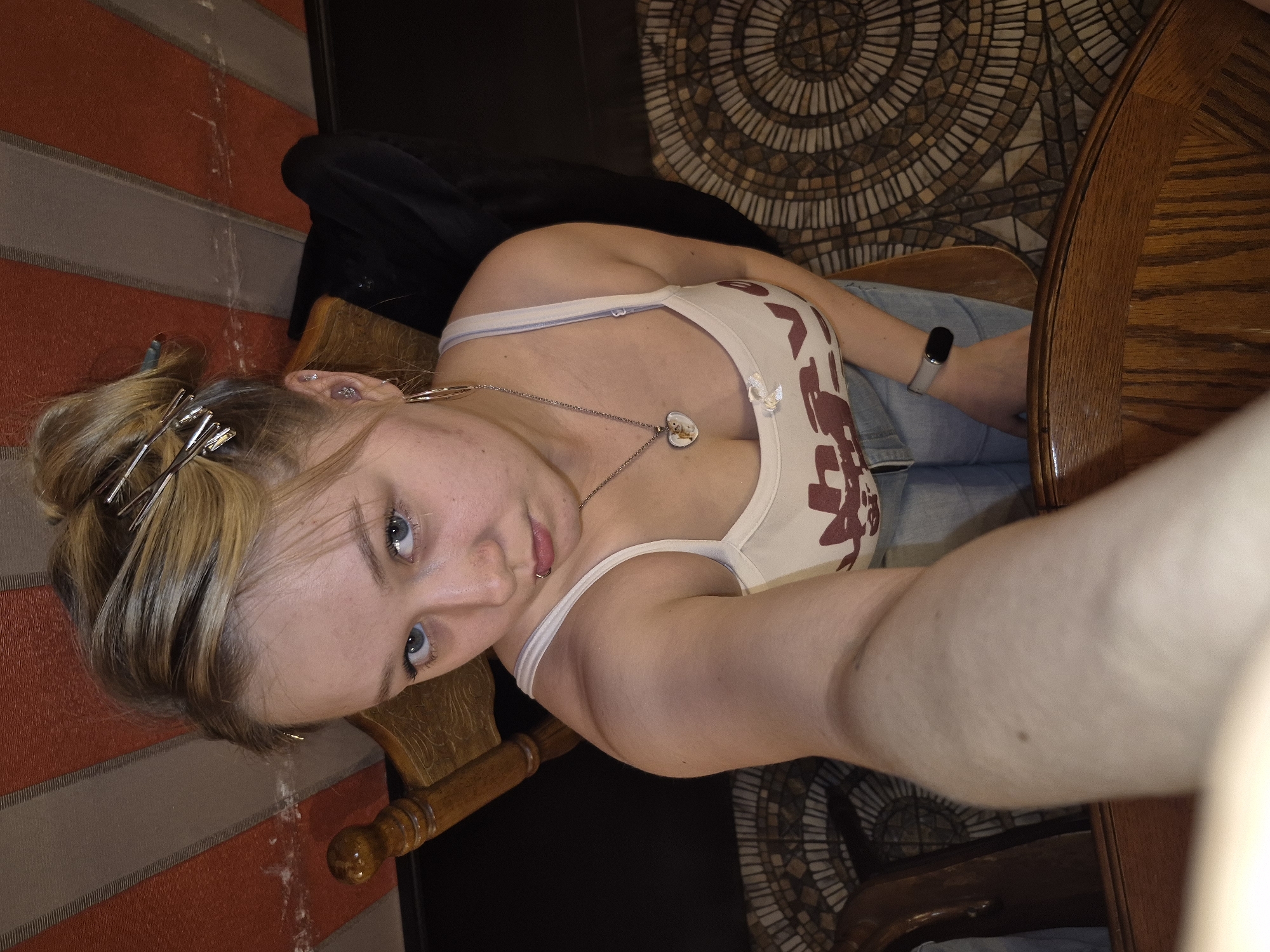 This is a first-person perspective photograph taken indoors, likely in a home or café, with the subject sitting at a wooden table. The flooring features a circular mosaic pattern, and there is a striped wall in the background. The subject, a young woman with blonde hair styled up and secured with metallic clips, is wearing a tank top with a graphic print and a necklace with a heart-shaped pendant. She has blue eyes and is looking directly at the camera with a neutral expression. A smartwatch is visible on her wrist. The lighting suggests it could be daytime. The photo appears to be a selfie, possibly taken by the subject herself.
