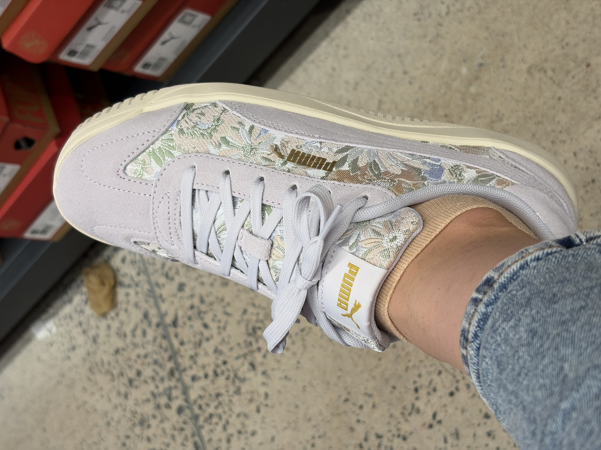 The image shows a close-up of a person's left foot wearing a light purple Puma sneaker with a floral pattern on the side. The sneaker has white laces and a cream-colored sole. A gold Puma logo is visible on the tongue of the shoe. The person is wearing light blue jeans and a tan ankle sock. The background shows a speckled floor and stacks of red Puma shoe boxes. The scene appears to be in a retail store.