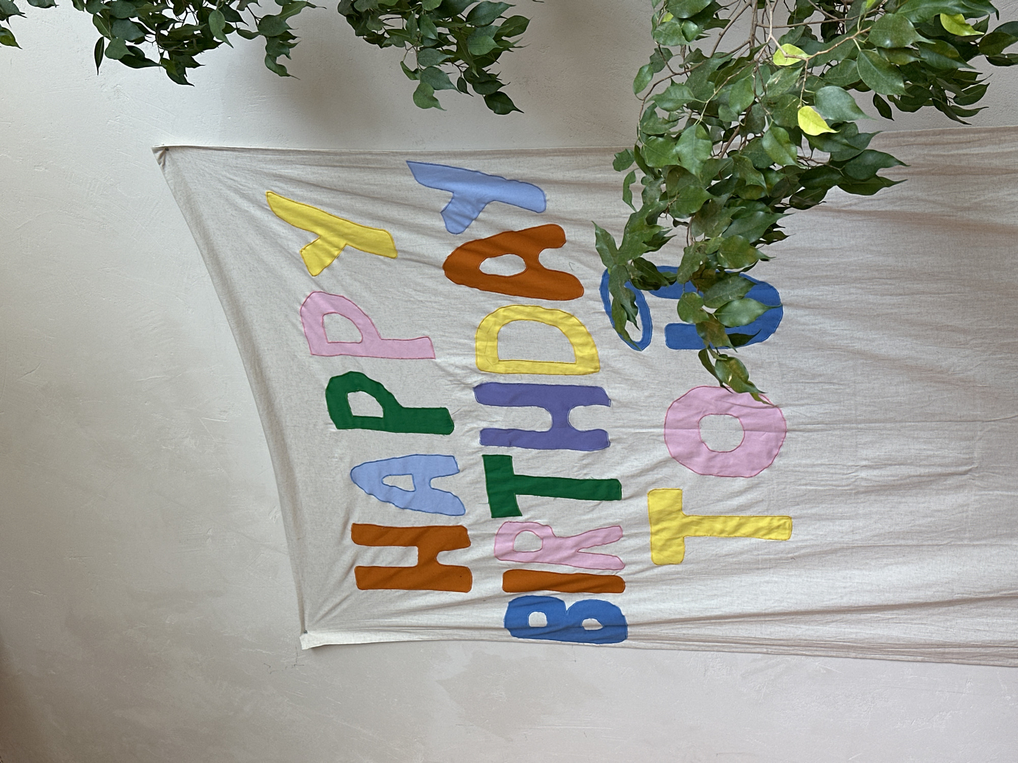 A horizontal photograph shows a "Happy Birthday to OT" banner hanging on a wall, partially obscured by hanging greenery. The banner is made of light beige fabric, and the letters are colorful and appliqued. The letters spell out "Happy Birthday to OT" vertically. The letters are various colors including yellow, pink, green, blue, and brown. The greenery is a leafy branch, and it hangs in the upper center and right of the frame. The wall is a light color. It cannot be determined whether this image was taken in the city of Batumi, Georgia from the provided information.