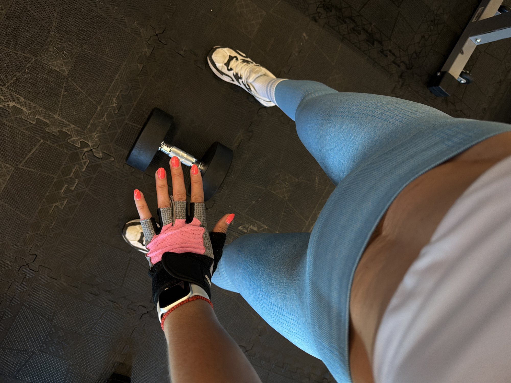The image captures a first-person, high-angle perspective of a person in a gym or home workout setting, located in Birobidzhan, Russia. The individual is wearing light blue, textured athletic leggings and a white top, with their midsection visible between the garments. On their feet are white sneakers with black patterns and white socks.
The person's left hand, adorned with bright coral-colored nail polish, wears a pink and black weightlifting glove. A silver smartwatch with a red braided band is visible on their left wrist. A black dumbbell rests on the floor beside their hand.
The setting consists of dark grey, interlocking rubber gym floor mats featuring various textured patterns, one of which faintly displays the letters "MB." In the upper right corner, part of a silver metal exercise machine frame, possibly a weight bench or rack, can be seen. The overall scene suggests a moment during or immediately before a workout session in an indoor environment.