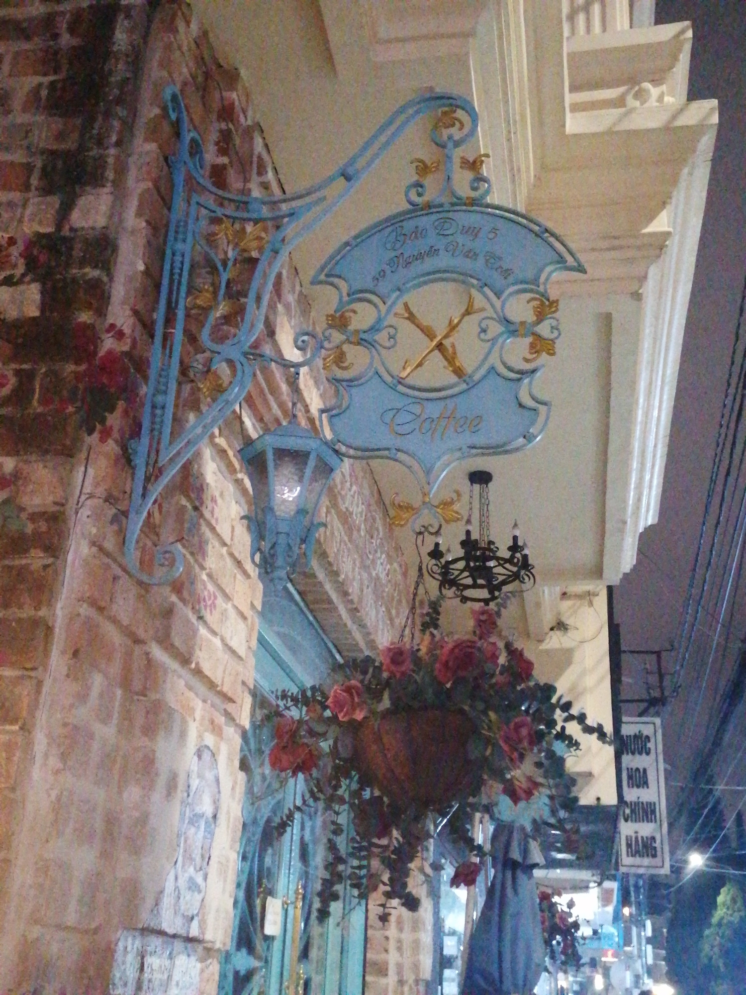 This eye-level shot captures the ornate exterior of a coffee shop or cafe at night in Da Lat, Vietnam. A prominent, elaborately designed sign, colored light blue with gold accents, hangs from a wrought-iron bracket attached to a brick wall on the left. The sign features scrollwork and leaf motifs, displaying the text "Bảo Duy 5" and "30 Nguyễn Văn Cừ" at the top, and "Coffee" in a script font at the bottom. In the center of the sign, two crossed branches or sticks are depicted.
Below the main sign, a matching blue ornate lantern hangs, providing illumination. Further down and to the right, a large hanging basket overflows with vibrant red roses and green foliage, adding a decorative element to the facade. A black wrought-iron chandelier is also visible, hanging slightly behind the flower basket.
The building on the left exhibits a brick facade, while the adjacent building on the right has a lighter-colored, smoother surface with classical architectural details such as pillars and cornices. The dark background suggests it is evening or night, with artificial lights illuminating the scene. In the distance, against the night sky, power lines crisscross, and a vertical white sign with Vietnamese text is partially visible, reading "NƯỚC HOA CHÍNH HÃNG," which translates to "Genuine Perfume." The scene conveys a charming and decorated street-side establishment.