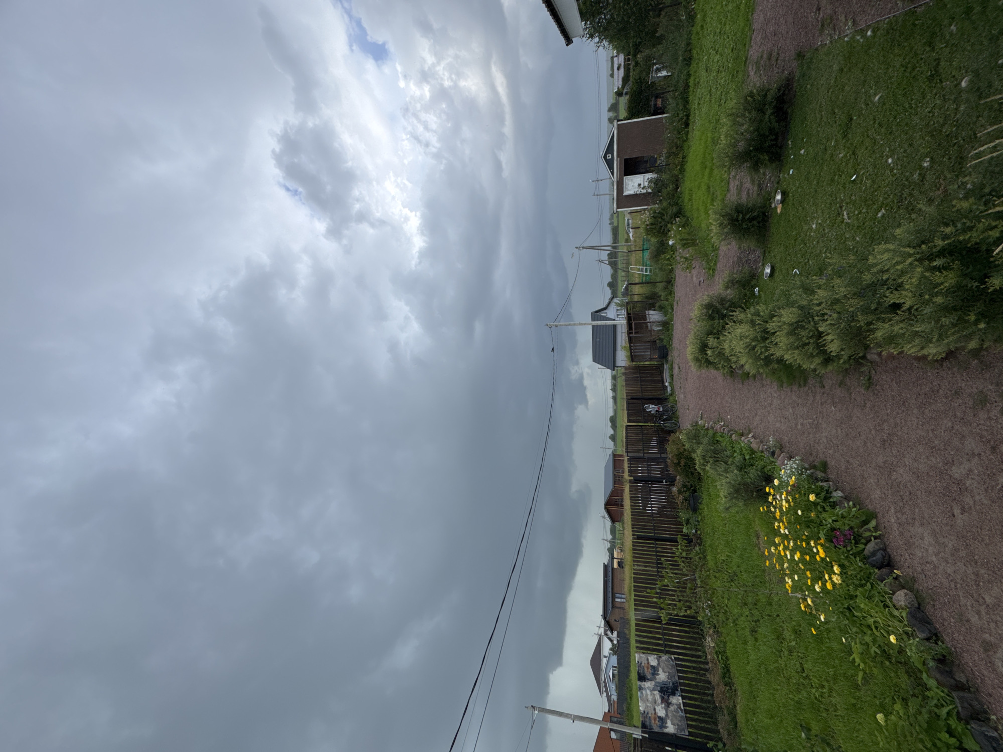 The image captures a view of a rural residential area in Kempelevo, Russia, under a heavily overcast sky, suggesting impending rain or storm. A wooden fence runs horizontally across the mid-ground, separating a grassy yard with a gravel pathway from a further expanse of greenery and several small houses. The pathway is lined with manicured bushes and a flowerbed containing yellow and purple flowers. In the background, across the fence, various structures are visible, including houses, sheds, and utility poles with overhead wires. A bicycle is parked against the fence on the right side. The sky dominates the upper portion of the image, filled with dark, heavy clouds. The overall impression is one of a quiet, somewhat gloomy, rural setting.