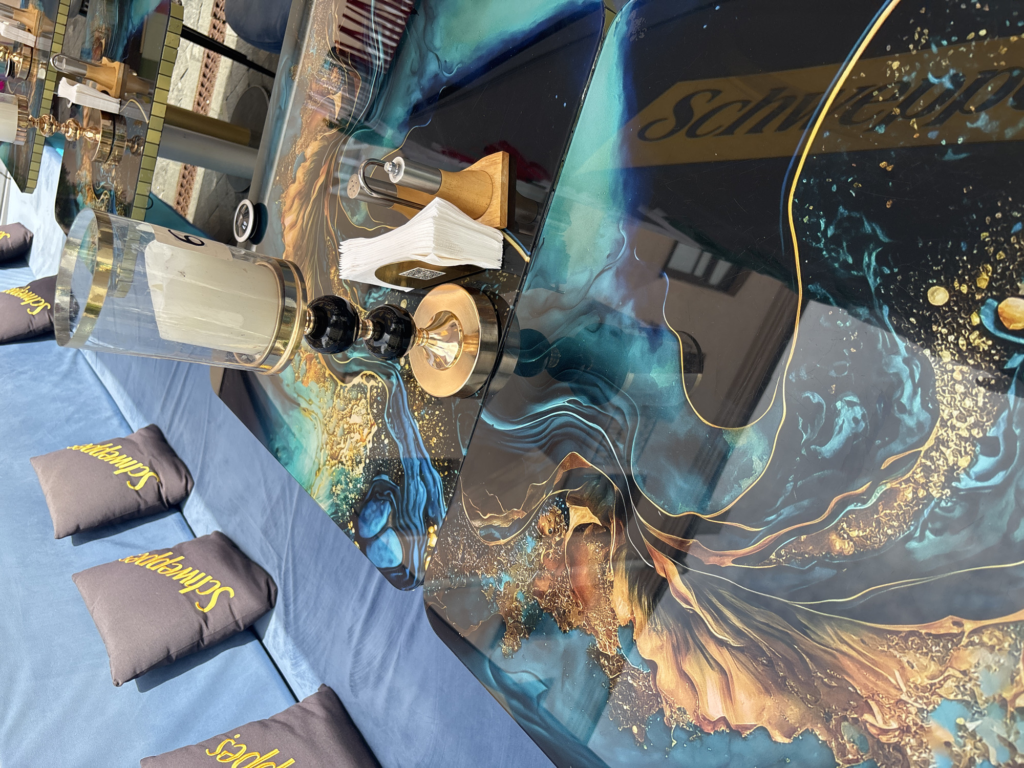 The image shows a close-up view of an outdoor seating area with decorative tables and seating. The seating is upholstered in blue fabric, and there are three dark gray throw pillows with yellow "Schweppes" branding visible on the couch. The tables have a glossy, resin-like surface with a marbled pattern featuring shades of teal, black, gold, and brown. On one of the tables, a glass hurricane lamp with a white candle is present, alongside a gold-toned candle holder with black accents, a wooden napkin holder with white napkins, and a salt and pepper shaker set. Another table in the background also displays a similar marbled surface and appears to be set up for dining. The overall ambiance suggests a stylish cafe or restaurant setting. The background is blurred but hints at an urban environment with buildings and pavement.