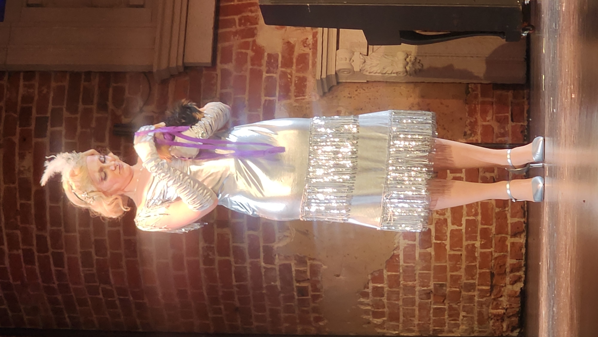 A blonde woman stands center frame on a wooden stage in front of a brick wall. She is wearing a silver, sleeveless dress with fringe at the hem, long silver gloves, and silver heeled shoes. She has a feather in her hair and is holding a puppet with purple ribbons. A dark, upright object, possibly a piano, is to the right of the frame. The lighting suggests this is a theatrical performance. The image does not provide any information about the location being Moscow, Russia.