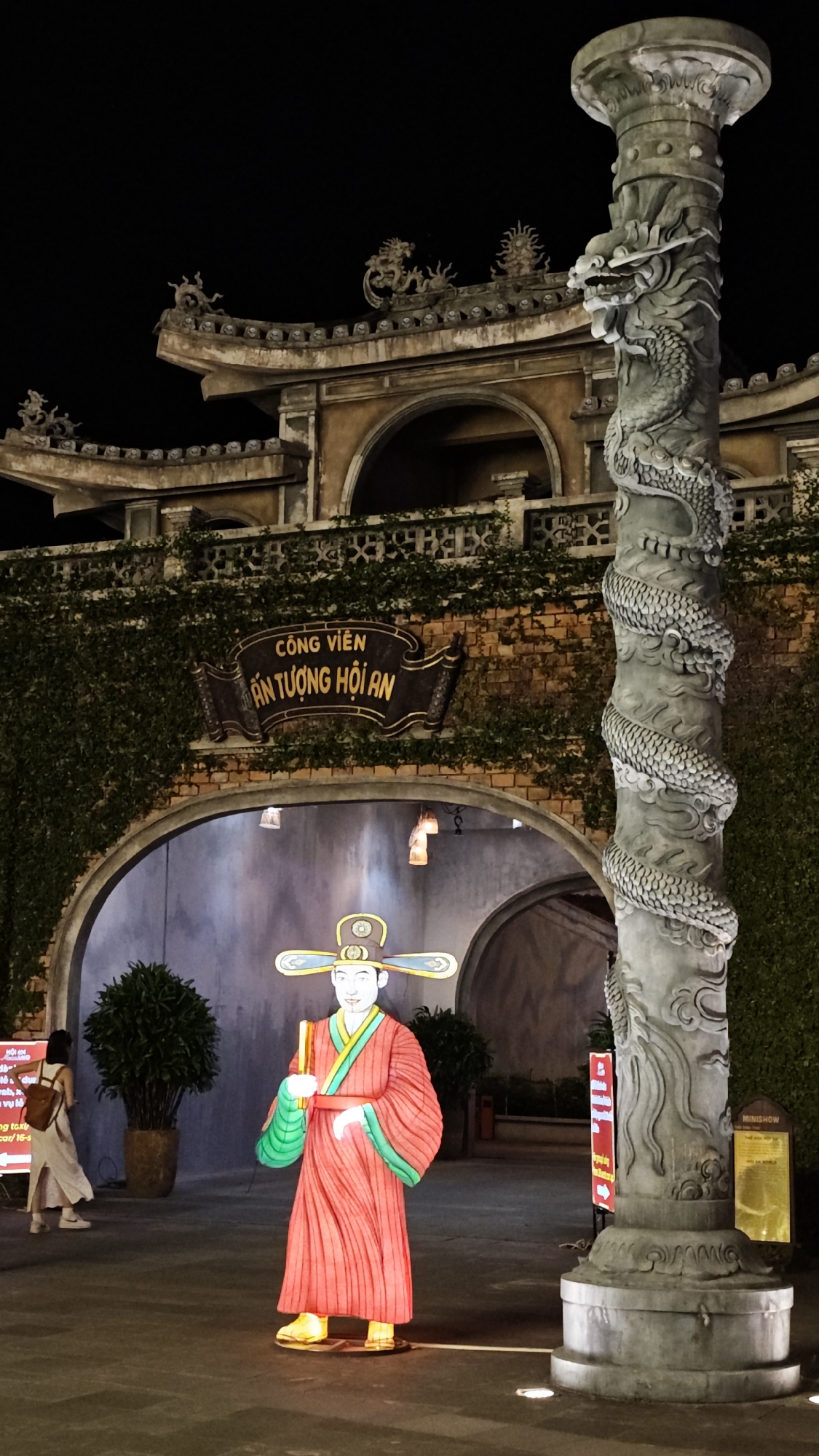 The image depicts a nighttime scene in Hội An Ward, Vietnam, showcasing cultural elements. In the foreground, there's a lit-up figure resembling a man in traditional Vietnamese attire. Next to it stands a grey stone pillar intricately carved with a dragon.
Behind them is an arched entranceway, above which a sign reads "CÔNG VIÊN ẤN TƯỢNG HỘI AN", indicating the location as a park or attraction in Hội An. The structure is partially covered in greenery, and its upper portion is decorated with dragon motifs. A person is walking in the background towards the archway. The overall lighting suggests an evening setting.