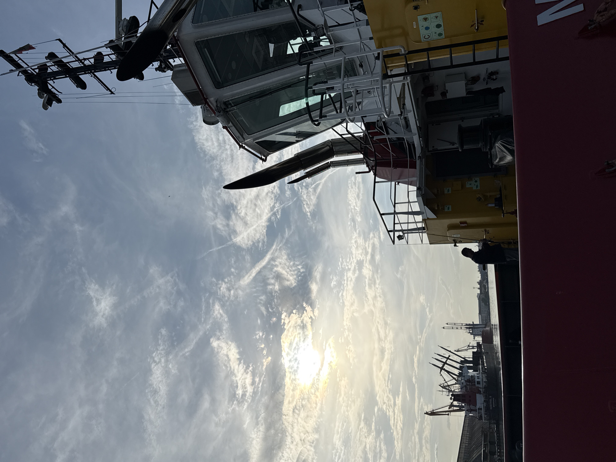The image captures a scene in Burgas, Bulgaria, with a ship as the prominent subject. A smaller ship is visible alongside a taller crane on the harbor. The sky is partly cloudy with the sun shining through the clouds. A silhouetted person stands on the deck of the ship, near a red structure. A cabin with multiple windows and antennas on top of the ship is in the top part of the frame.