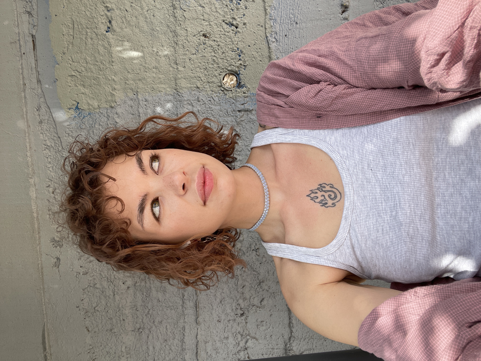 A young woman with curly brown hair is positioned on a textured gray surface. She wears a white tank top, a pink checkered shirt, and a diamond-like choker necklace. A small tattoo is visible on her upper chest. She is looking upwards, and her face is slightly tilted. The background includes a light gray concrete wall. Based on the visual context, the location is Rafailovići, Montenegro.