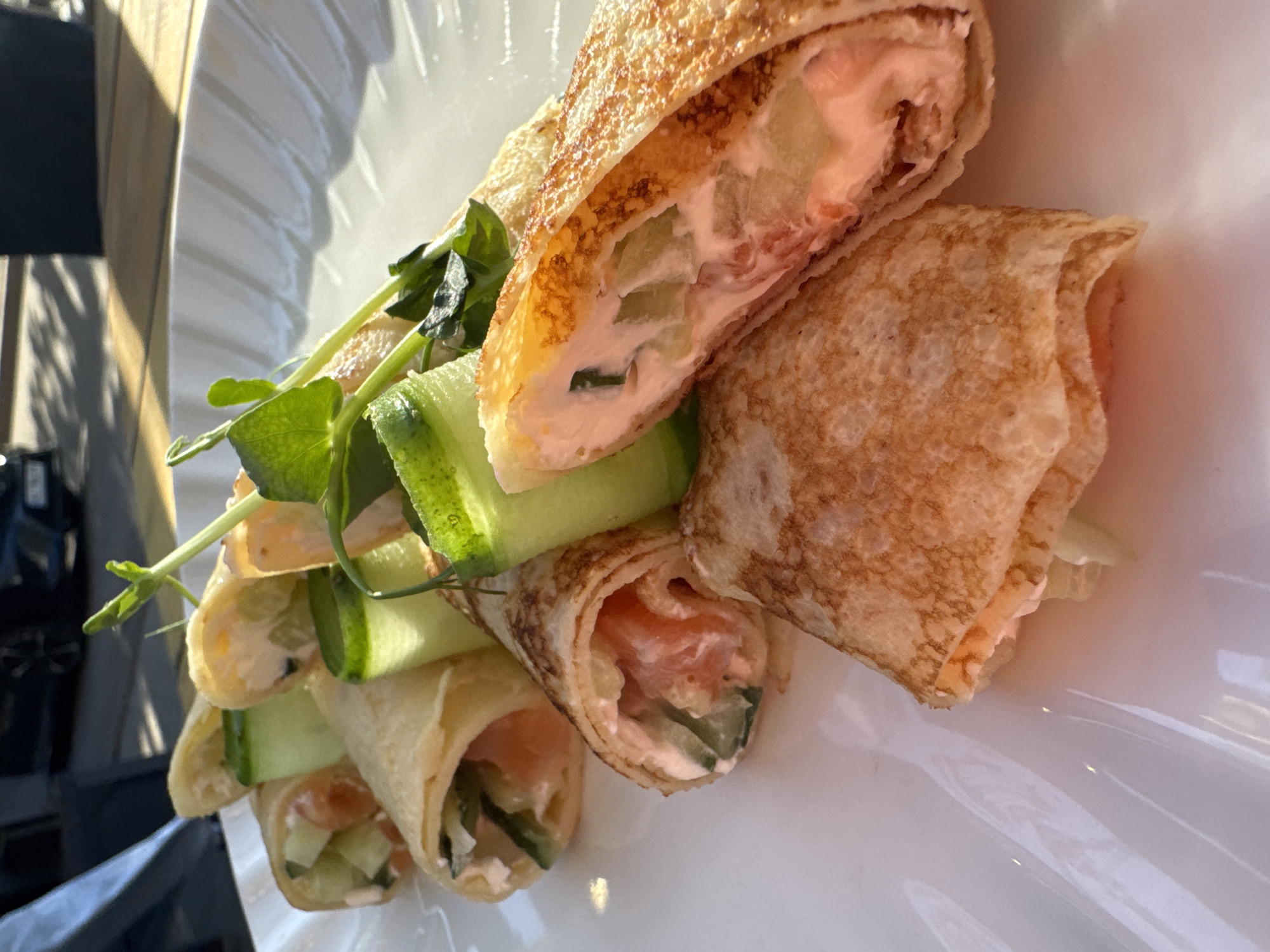 A close-up shot shows several rolled crepes filled with what appears to be salmon, cucumber, and a creamy spread, arranged on a white plate. The crepes are stacked and garnished with green sprouts. The background is blurry, but indicates a table setting near a window with natural light. The location, Kryvyi Rih, Ukraine, is not evident from the image itself.