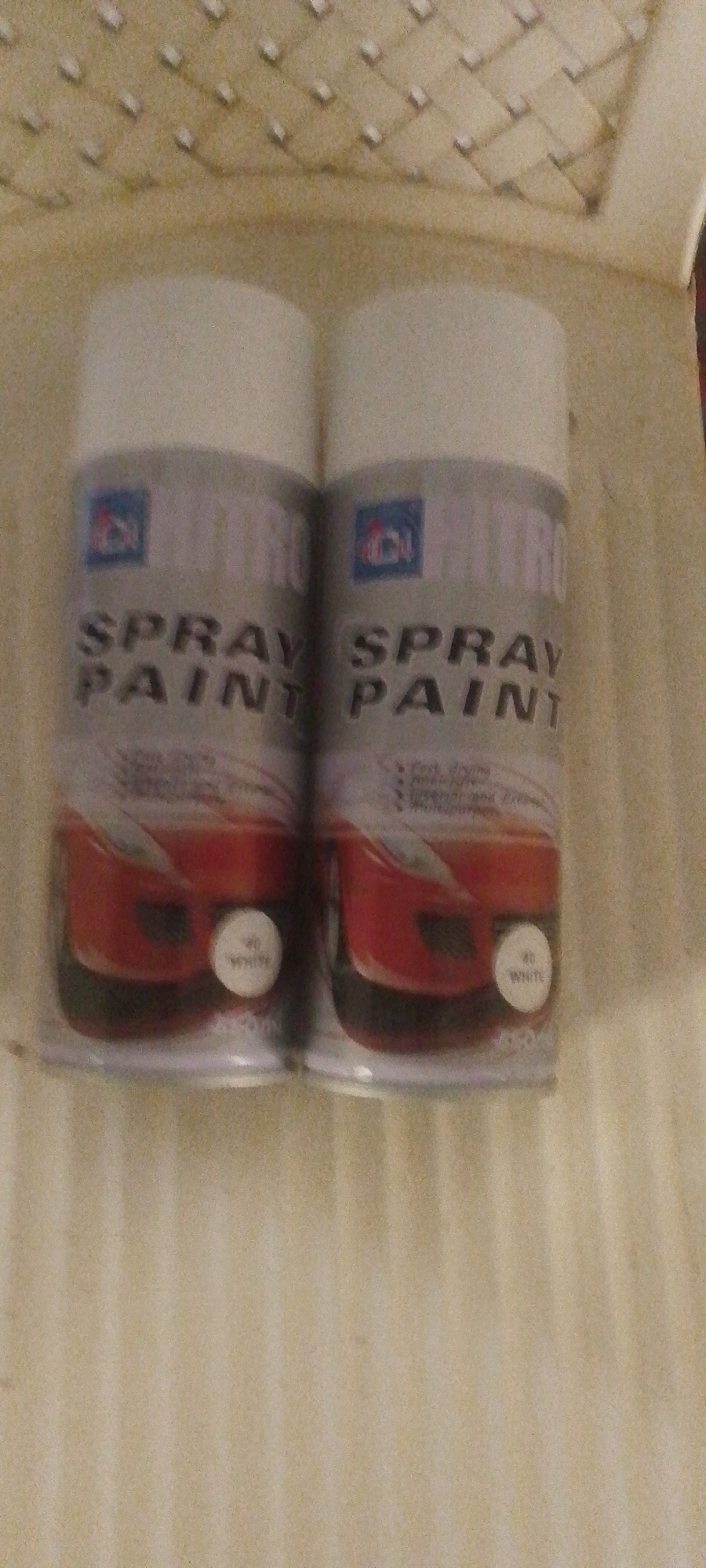 Two cans of white spray paint are placed side-by-side on a textured, light-colored surface. The cans are white with silver lids and have labels that read "NITRO SPRAY PAINT" in large, bold letters. Below this text, there is an image of a red car. A small circular label on each can displays the number "40" and the word "WHITE". The background appears to be a textured surface with a repeating pattern of raised squares. The overall lighting suggests an indoor setting. The image does not depict any recognizable landmarks or elements that would allow for specific location identification within Jalingo, Nigeria, or any other city.